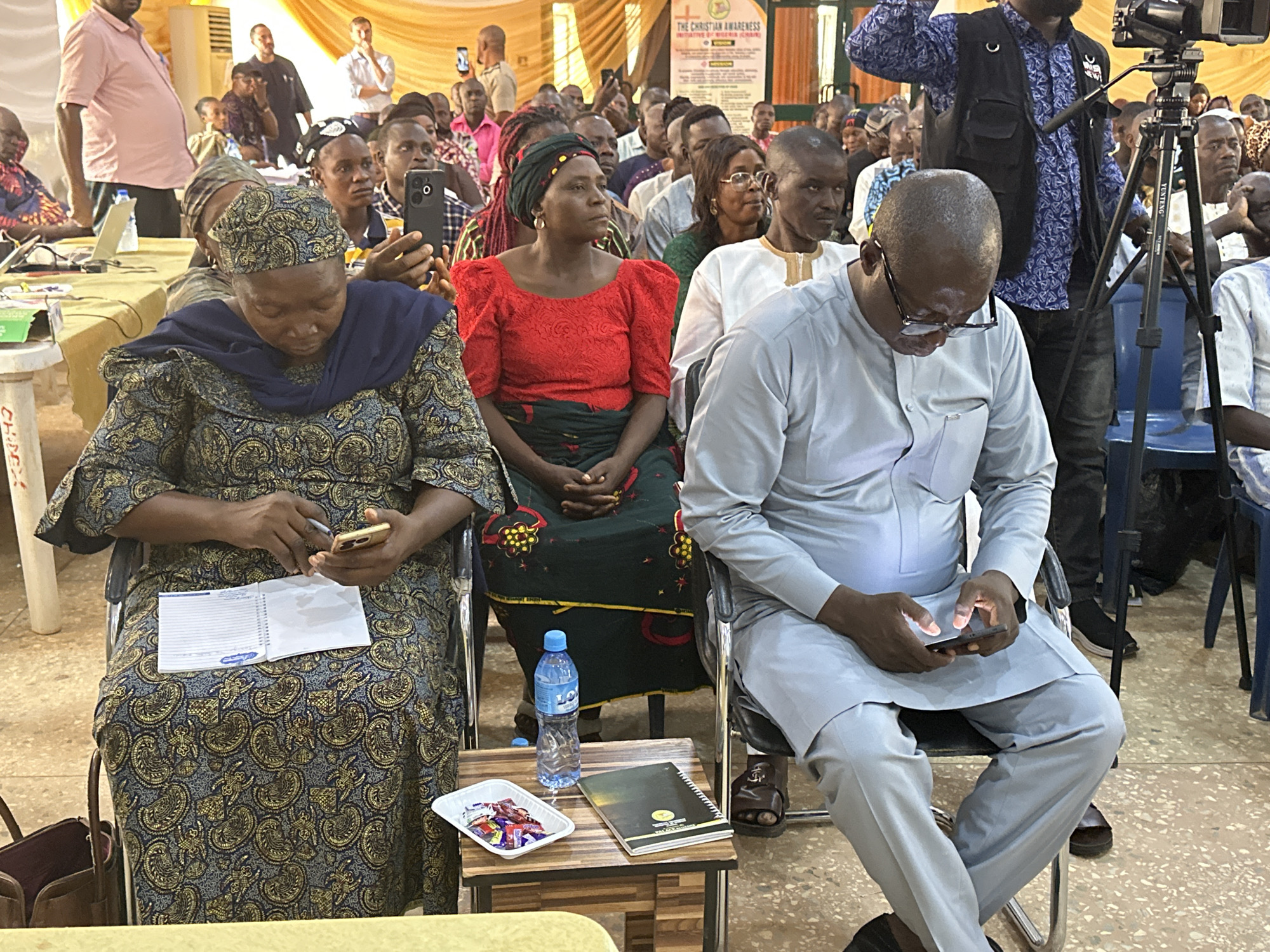 A medium shot captures a diverse group of people seated in an auditorium, seemingly attending a conference or meeting. The primary subjects are three individuals in the foreground: a woman in a blue and gold patterned dress and headwrap is engrossed in her phone while holding a notebook. Beside her, a woman in a red blouse with puffed sleeves and a green and black patterned skirt sits with her hands clasped. To their right, a man in a light grey traditional attire focuses on his smartphone.
A small wooden table beside them holds a bottle of water, a dish of individually wrapped candies, and a notebook. Behind them, a densely packed audience fills the frame, many holding phones, suggesting active engagement with the event. A video camera mounted on a tripod stands to the right, indicating that the event is being recorded.
In the background, a banner with the words "THE CHRISTIAN AWARENESS" is visible. The room is lit with natural light filtering through unseen windows, supplemented by artificial lighting. The location is in the city of Unknown, Unknown.