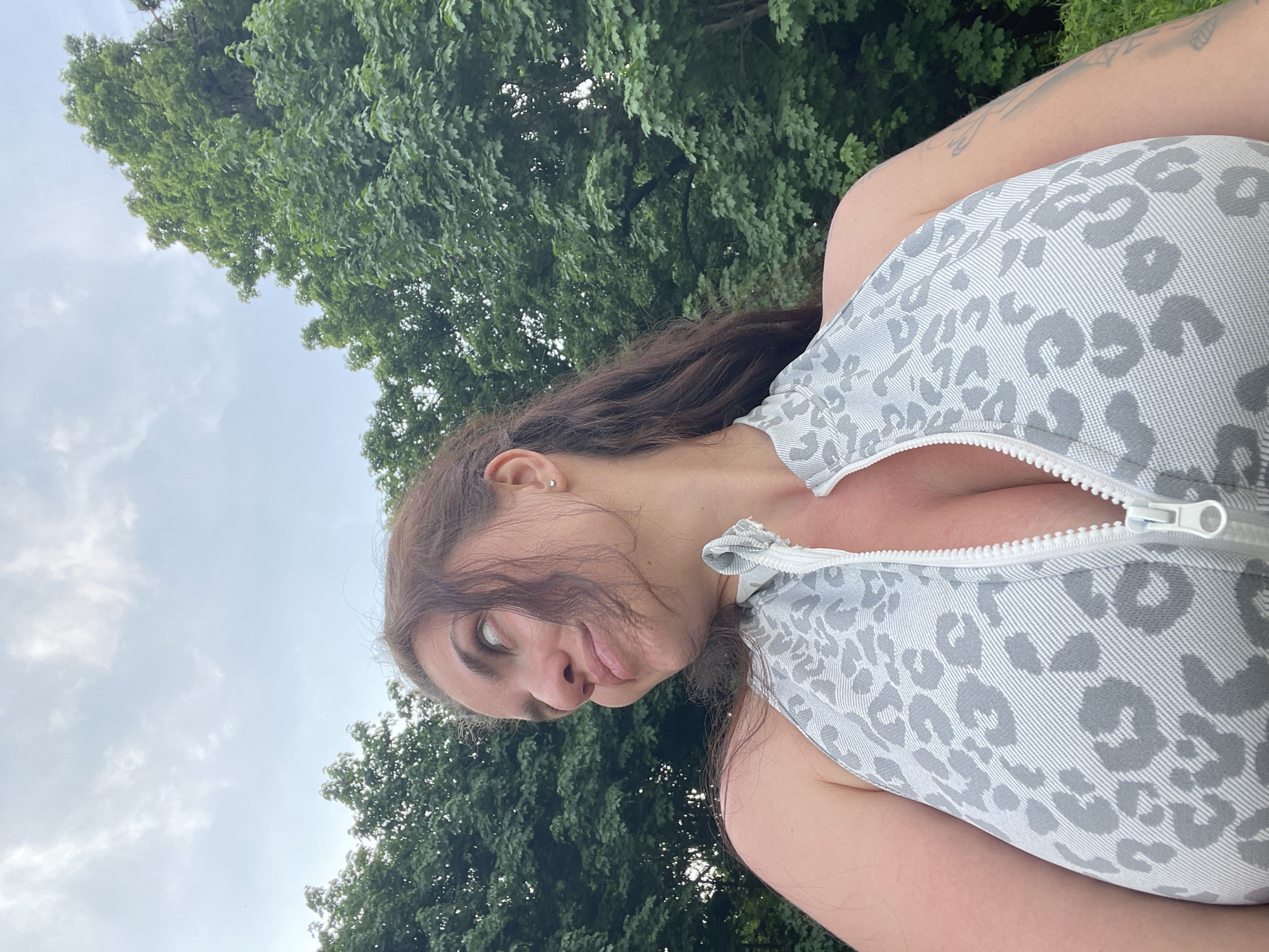 The image is a selfie taken outdoors, likely in a park or other green space. A woman is the main subject, posing with a slightly goofy expression; she is tilting her head to the side and sticking out her tongue. She has dark hair and fair skin, and she is wearing a gray and white leopard print garment with a zipper. Part of a tattoo is visible on her upper arm. In the background, there are trees with lush green foliage, and the sky is overcast with some patches of white clouds. The image does not include any information to indicate the location of Ostrov, Russia.