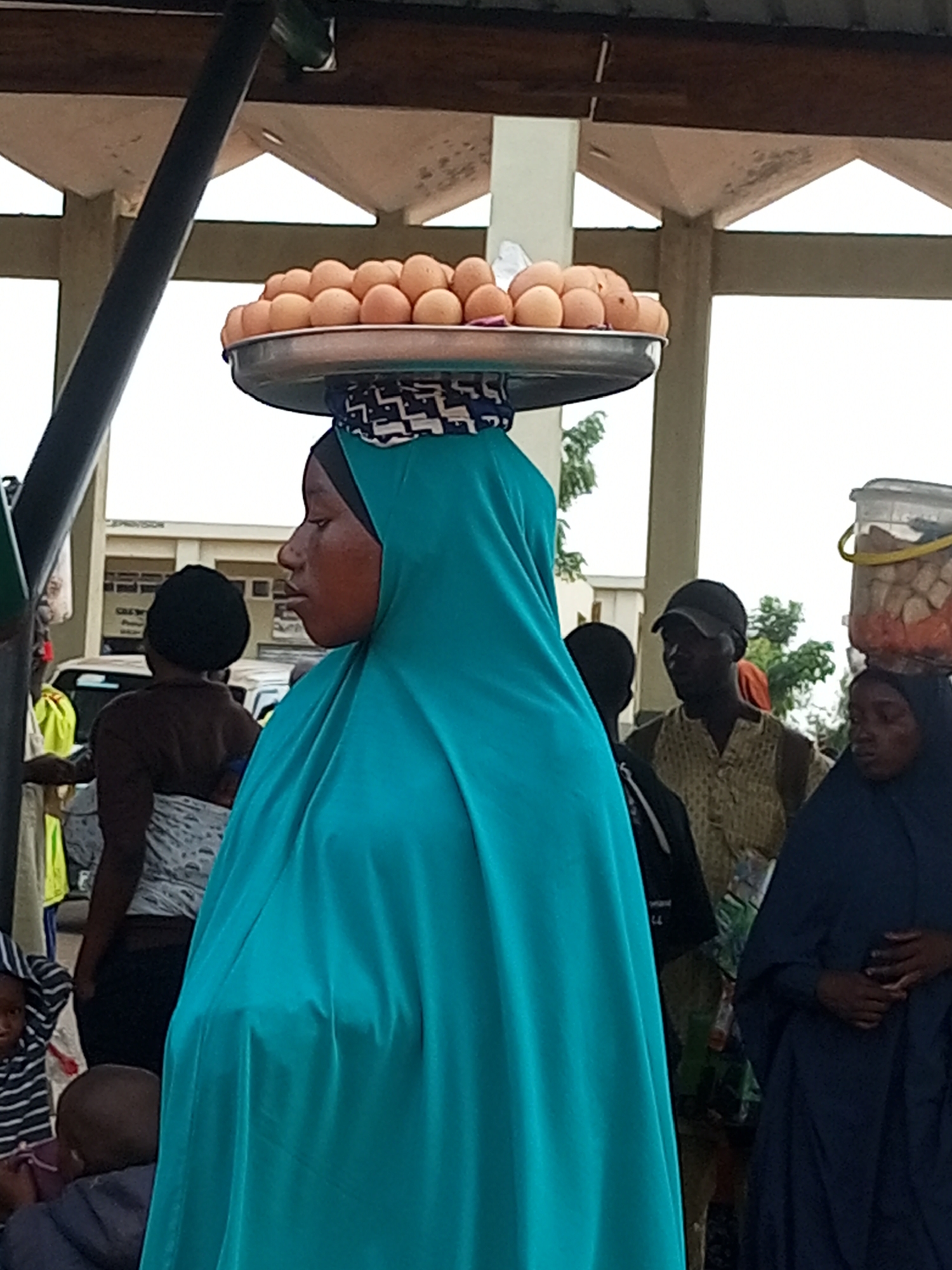 A street scene in Gombe, Nigeria, features a woman wearing a teal hijab and blue dress, balancing a large metal tray piled high with eggs on her head. She is standing in the foreground, facing right, with her mouth slightly open. Behind her, a woman in a dark hijab and dress stands to the right, also facing right, with her hands clasped. To the left of the main subject, another woman in a dark headscarf and patterned skirt faces away, carrying a child on her back. In the middle ground, a man wearing a patterned shirt and a dark baseball cap faces left. Further back, other people and vehicles are visible, and the environment appears to be an outdoor market or thoroughfare with a covered structure overhead.