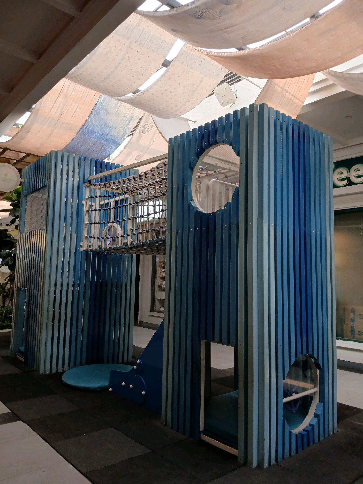 A modern blue wooden playground structure is centrally located on a black checkered mat floor. The structure is multi-level with various openings, including a circular tunnel-like entrance on the right and a bridge made of rope netting connecting two sections. The playground has a whimsical design with vertical wooden slats in varying shades of blue. Above the structure, a canopy of draped fabric, with subtle patterns, filters the light, creating a soft ambiance. In the background, a green sign with white lettering, possibly an advertisement, is visible. The overall setting suggests an indoor recreational space, likely within a shopping mall or public area in North Jakarta, Indonesia, designed for children's play and exploration.