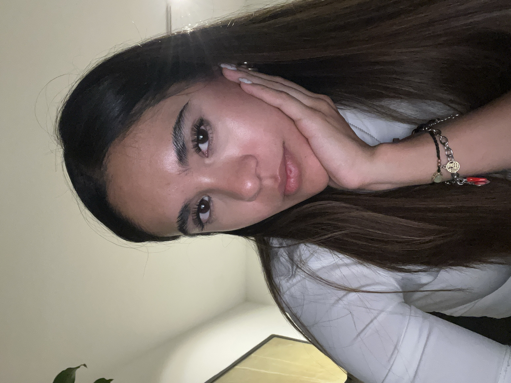 A young woman with long brown hair is posing indoors, resting her face in her hand. She is wearing a white long-sleeved shirt. A bracelet with charms is visible on her wrist. The background is a neutral, light-colored wall with a glimpse of a plant and a framed object. The image is oriented at an angle. There are no discernible indicators within the image to confirm the location as San Bernardino Tlaxcalancingo, Mexico.