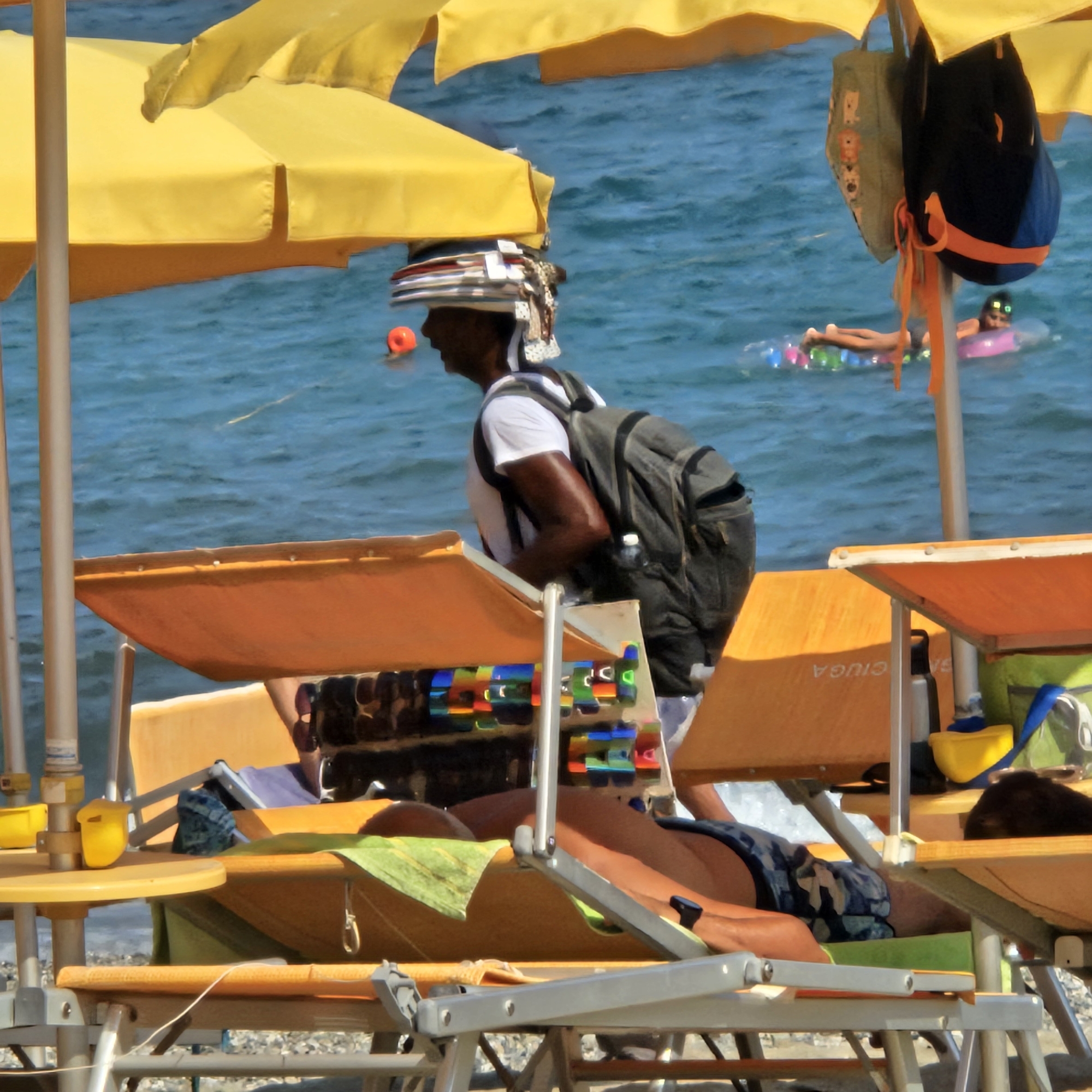 The image depicts a beach scene in Savona, Italy, focusing on beach chairs, umbrellas, and people enjoying the water. In the foreground, several orange beach chairs are visible, some with yellow cushions. Two yellow beach umbrellas provide shade. A person is seen wearing a backpack and a hat made of stacked items, walking past the chairs. In the background, the blue sea is calm with small waves. A person is floating on a colorful inflatable raft in the water. An orange buoy is also visible in the water. The overall atmosphere suggests a sunny day at the beach.