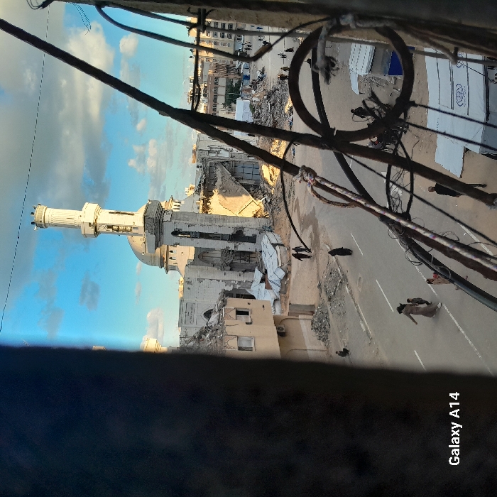 The image depicts an extensively damaged urban area in Khan Yunis, Palestinian Territory, captured from an elevated viewpoint. In the background, a large mosque with a prominent minaret and dome stands severely damaged, with significant portions of its walls and roof collapsed into extensive rubble. Surrounding buildings also display substantial destruction. A paved street traverses the middle ground, on which multiple individuals are walking: a person in a light-colored garment walking with a child, another person in dark attire further down the road, and a figure crouching near a pile of debris on the left. In the mid-right, several white tents, one clearly branded "UNRWA," are situated amidst further ruins, with additional individuals present nearby. The foreground is partially obscured by metal bars and wires. The scene is illuminated by natural light, casting long shadows under a partly cloudy sky.