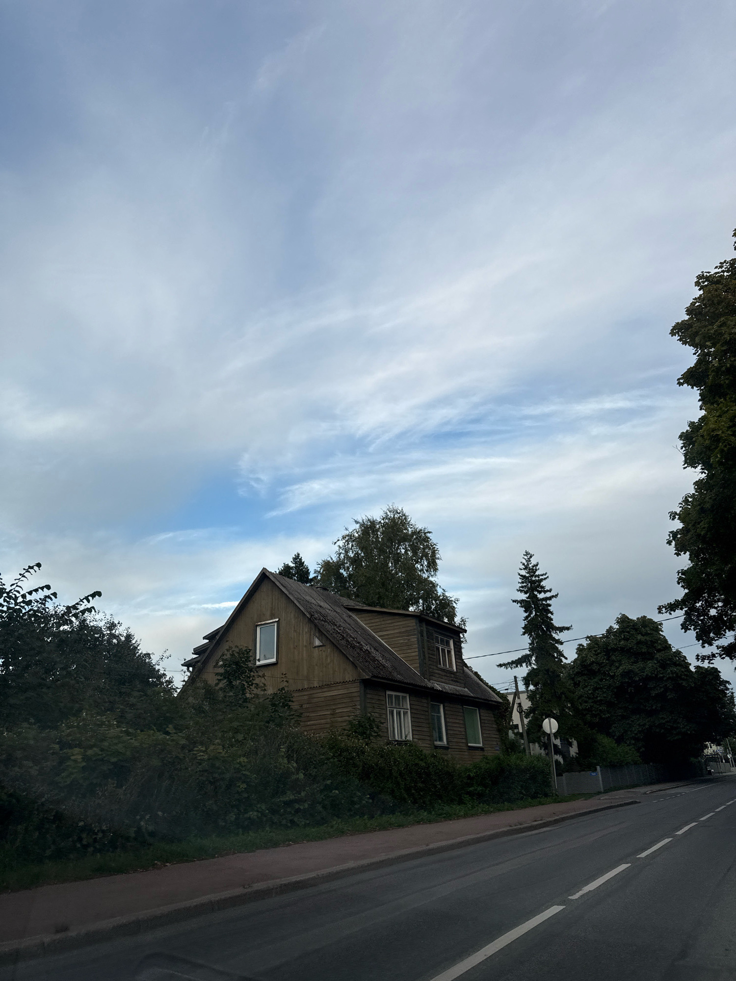 The image depicts a two-story wooden house partially obscured by dense foliage, situated along a paved road in Tallinn, Estonia. The house has a dark, weathered wood exterior with a steeply pitched roof covered in dark shingles. Several windows are visible, including dormer windows on the upper level. The surrounding area is characterized by lush greenery, with tall trees and bushes encroaching on the property and lining the street. The road in the foreground is asphalt with white lane markings and a paved sidewalk. The sky above is partly cloudy with wispy cirrus clouds against a pale blue backdrop. The scene appears to be captured from a low angle, possibly from a vehicle, given the perspective of the road. The overall impression is that of a residential area on the outskirts of the city, with a slightly overgrown and rustic aesthetic.