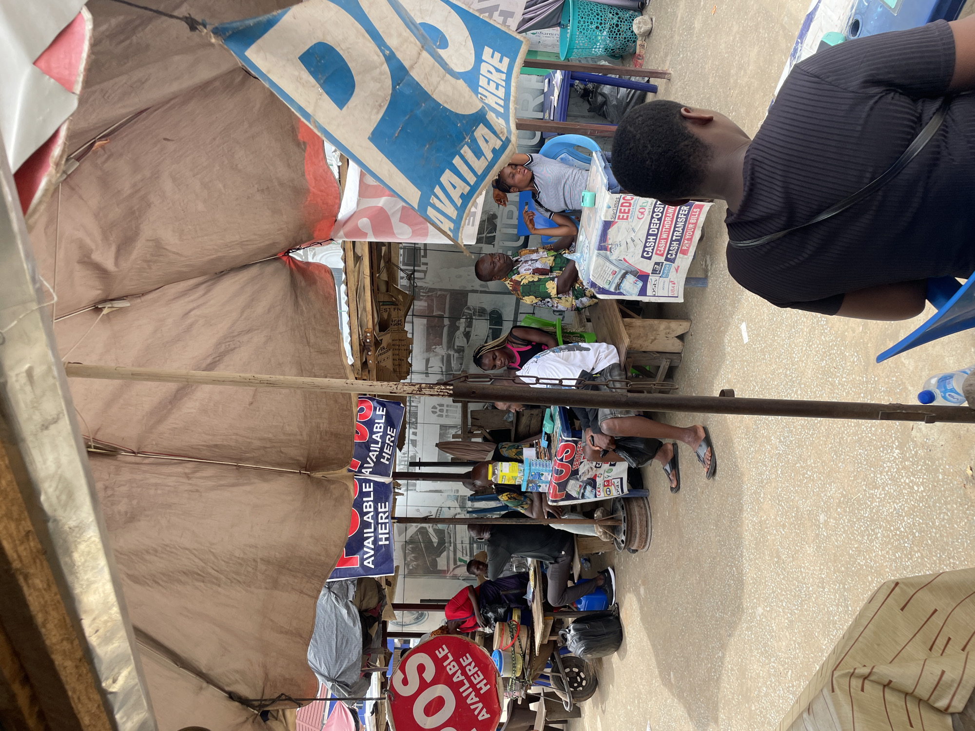 The image depicts a bustling outdoor scene in Onitsha, Nigeria. Several individuals are seated under makeshift shelters or tents, some engaging in transactions or conversations. Banners and signs advertising services like DStv, StarTimes, and various financial transactions such as cash deposits and withdrawals are visible. One sign features a red circle with the letters "SOS." The environment includes assorted items, such as plastic baskets, water bottles, and what appears to be equipment used for business transactions. The scene is captured from an eye-level perspective.