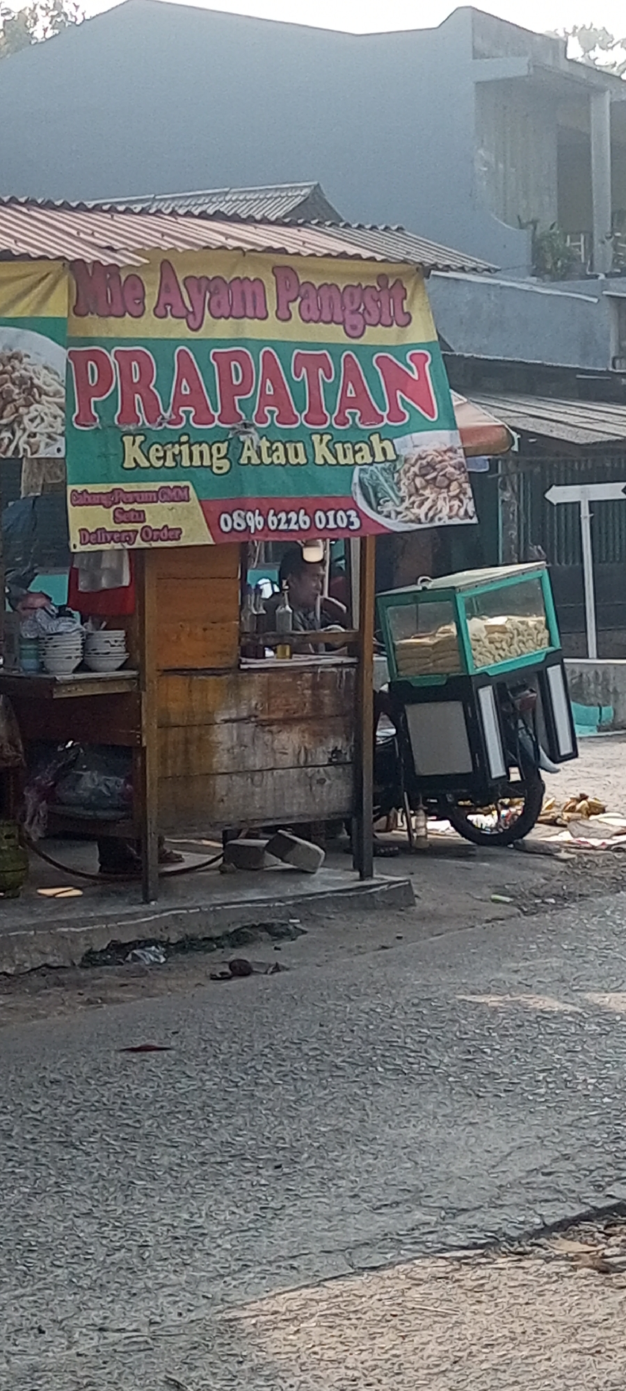 An outdoor food stall in Setu, Indonesia, displays a banner that reads "Mie Ayam Pangsit PRAPATAN - Kering Atau Kuah". The stall appears to be set up on the side of a road, with a wooden counter and a glass display case on wheels filled with food items. A person is visible behind the counter, seemingly attending to the stall. The background shows a building and some foliage. The ground in front of the stall is paved, with some debris scattered around. The overall scene depicts a typical street food vendor in an urban or semi-urban Indonesian setting.