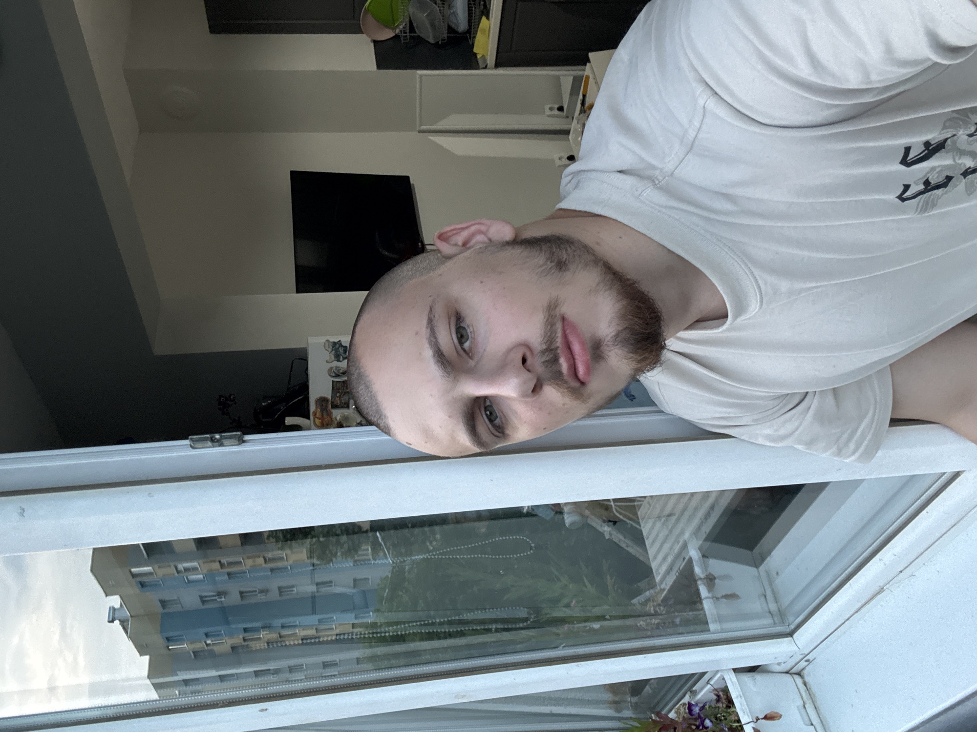 A man with light skin, short hair, and a beard is looking out an open window. He is wearing a light-colored t-shirt. The view through the window includes a building complex reflected on the glass. The window frame is white. Based on visible characteristics, the location is Stara Zagora, Bulgaria.