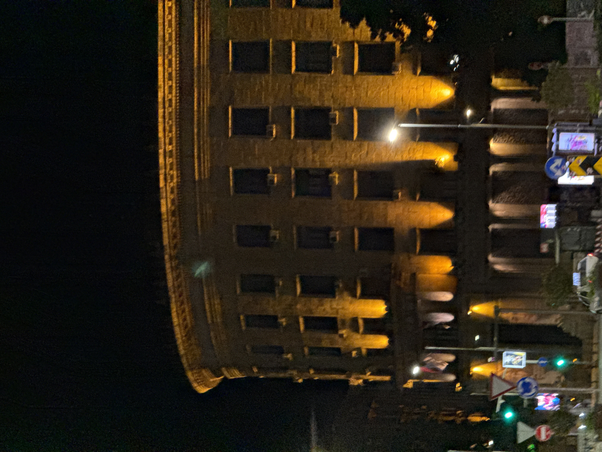 The image shows the night view of the building of the Main Post Office in Belgrade, Serbia. The building is illuminated with yellow light and has a curved facade. In the right part of the image, there are street lights and traffic signs. The left part of the image is mostly dark.