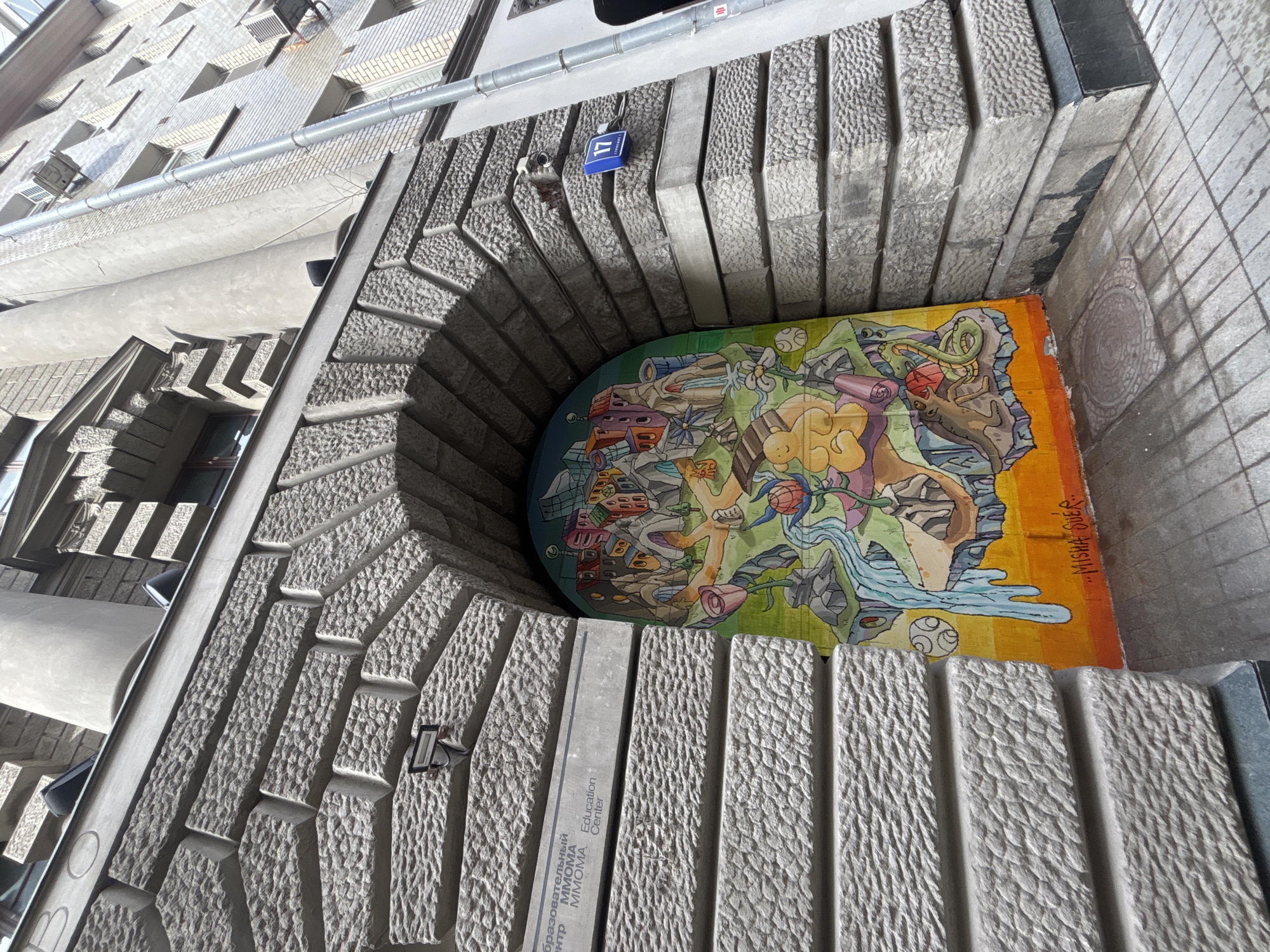 The image is a low-angle shot of an architectural feature in Moscow, Russia, specifically at the MMOMA Education Center. It features a mural or art installation displayed within a recessed archway of a building. The archway is constructed of light gray stone or concrete blocks with a rough, textured surface. A blue sign with the number "17" is affixed to the upper portion of the arch. The mural is multicolored with an abstract, somewhat surreal composition featuring elements of urban landscapes, natural forms, and cartoonish figures. Part of the mural has a signature "MISHA SUER" visible. The surrounding building architecture includes windows and ledges of a similar stone or concrete material. The sky is not visible, but lighting suggests it is daytime.
