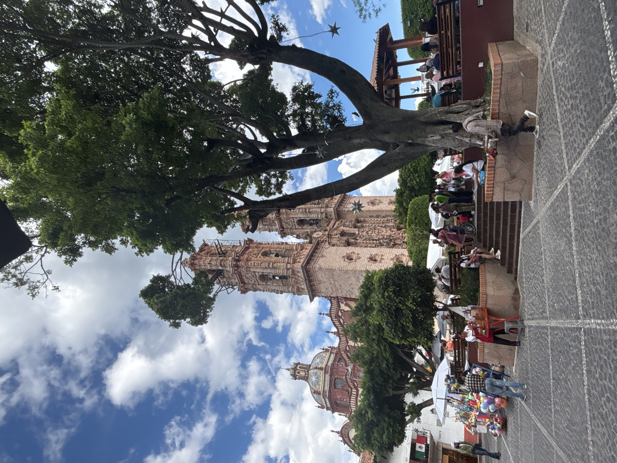 In Taxco, Mexico, a sunny day unfolds in a plaza dominated by the striking architecture of the Santa Prisca Church. The church, with its ornate pink stone facade, two bell towers, and a large, colorful dome, stands majestically in the background. A large, mature tree with spreading branches occupies the foreground, its leaves casting dappled shadows on the cobblestone plaza.
Under the tree, a small gazebo with a red tile roof provides shade for people seated on benches. Star-shaped decorations hang from the tree's branches. The plaza is bustling with activity. To the left, a vendor is selling a colorful array of balloons and other items from a stand beneath a white umbrella. A man in a plaid shirt and jeans is looking at the merchandise. Further to the left, a man in a green jacket stands near some flags.
In front of the church, a set of stone steps leads up to a seating area. People are gathered here, some sitting on benches, others standing. A woman in a red apron is tending to a small red table displaying food items. Several individuals are visible on the steps and in the seating area, engaged in conversation or resting.
The cobblestone ground is a textured surface with a pattern of intersecting white lines. The sky is a vibrant blue, dotted with fluffy white clouds. The scene captures a lively moment in a historic Mexican town, with the iconic Santa Prisca Church serving as a stunning backdrop to everyday life.