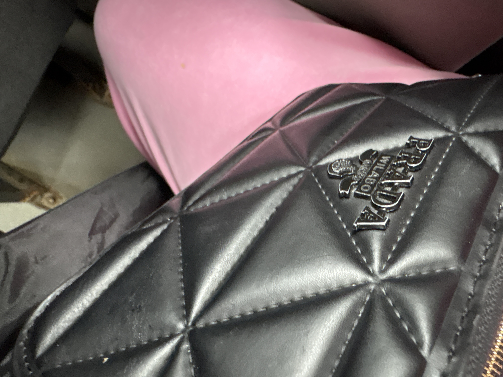 The image shows a close-up of a black quilted handbag with a silver logo. The handbag is resting on a person wearing a pink garment. The background is blurred and appears to be a dark, possibly upholstered surface. The image is taken from a high angle, looking down at the handbag. There is no visible indication of the location in the city of Mahilyow, Belarus, within the image itself.