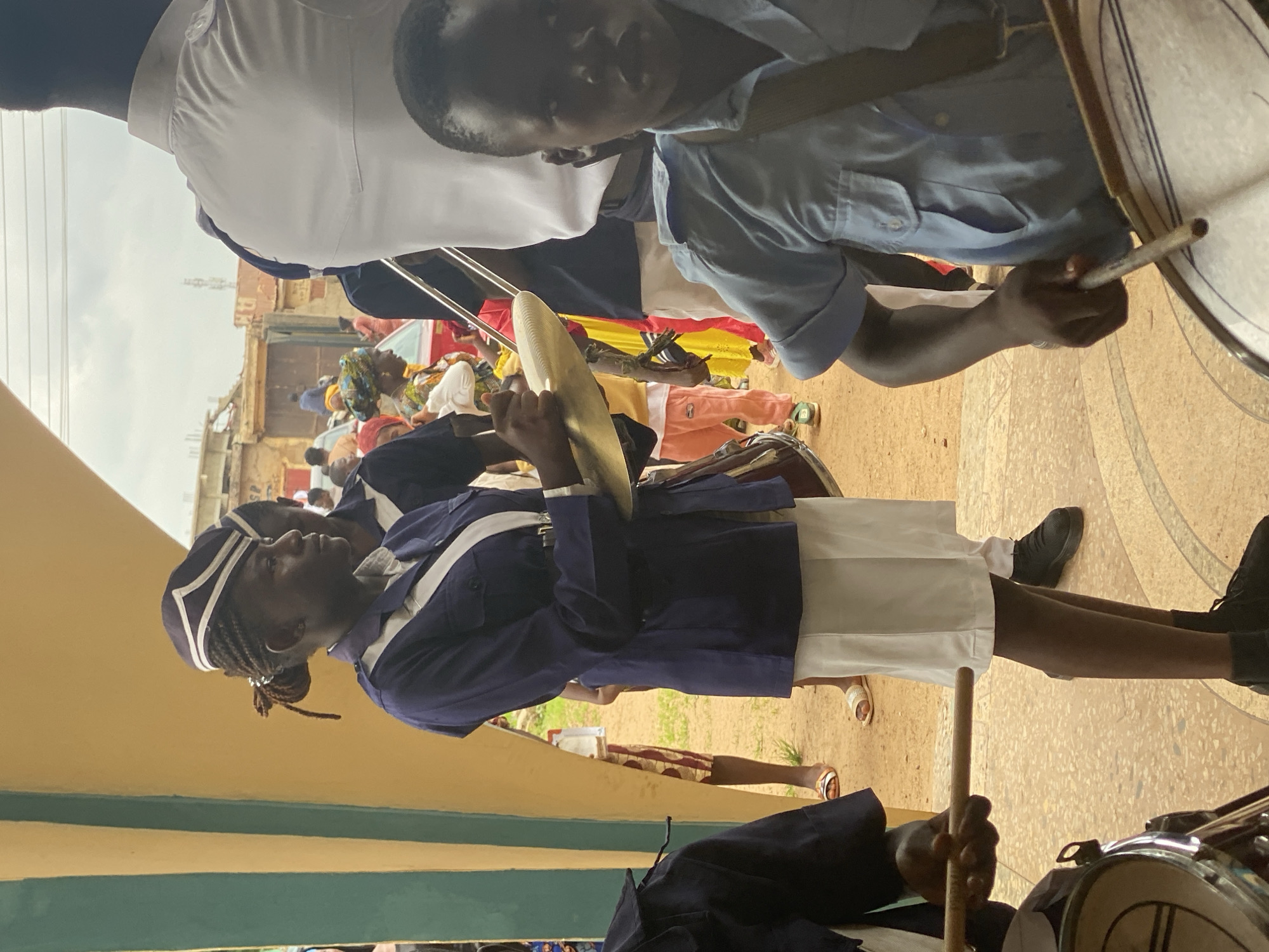 The image shows a procession in Kafanchan, Nigeria. A marching band is present, with a band member playing the cymbals prominently in the foreground. The band members are wearing blue and white uniforms. A drummer is partially visible, also wearing a blue and white uniform. Spectators are in the background, some wearing colorful head coverings. Buildings are visible in the distance. The location appears to be near or under a building overhang painted in yellow and green.