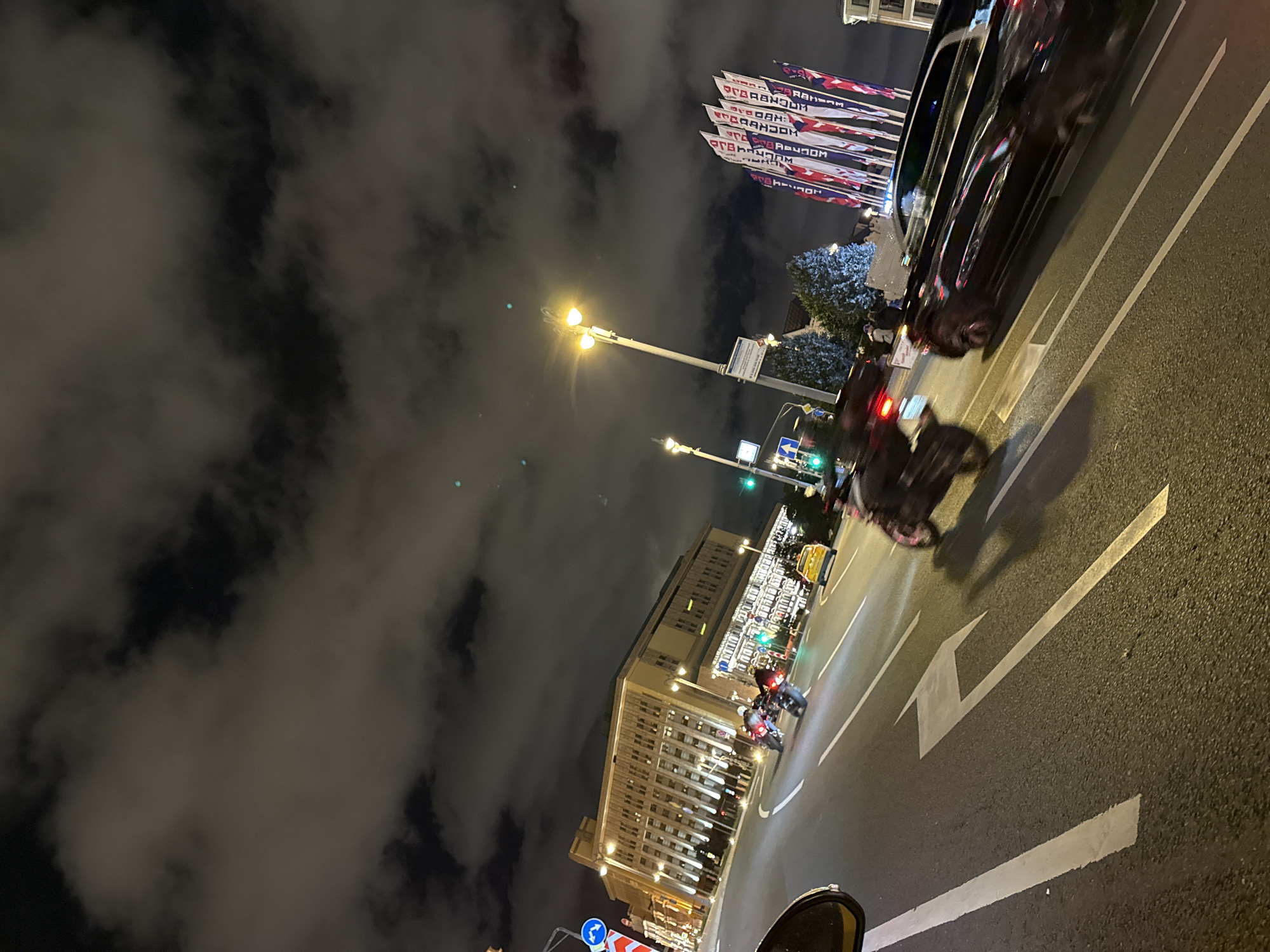 This is a night time, eye-level, medium shot captured from a moving vehicle looking towards a wide, paved road in Moscow, Russia. The road is marked with white lane lines, including a directional arrow indicating a left turn. On the left side of the road, there is a large, illuminated building with many windows. Further down the road, several motorcycles are seen, some appearing to be in motion. A yellow taxi is also visible on the right side of the road. In the background, there are streetlights and a line of flags, some of which bear Cyrillic text. The sky is dark and cloudy.