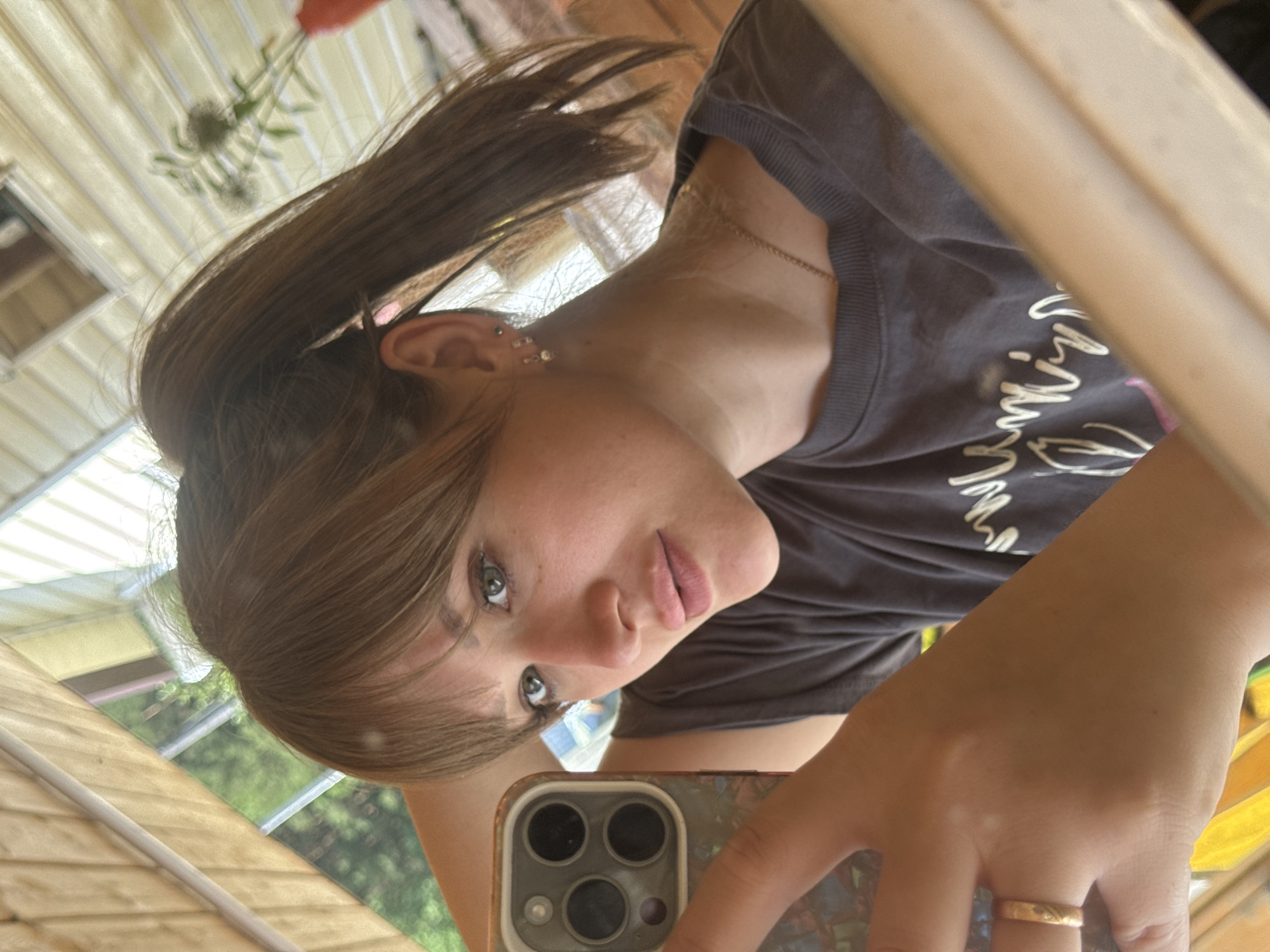The image depicts a female subject taking a mirror selfie indoors. Her medium brown hair is styled with bangs across the forehead and pulled back into a high ponytail. She has green eyes and her lips are coated with a light pink color. The visible left ear features three piercings: a small dark stud near the top and two small hoop earrings with clear stones. A thin, gold-colored chain necklace is worn around her neck. Her left hand, holding a mobile phone, displays a plain gold-colored band ring on the ring finger. The phone is an iPhone, identifiable by its triple-lens camera module, housed within a multi-colored case featuring graphic imagery. She is wearing a dark gray, ribbed crew-neck t-shirt with a faded light-colored cursive design on the front. The background reflected in the mirror through a window shows outdoor greenery, indicative of trees or bushes, and a light-colored building with horizontal paneling. A small, sparse plant in a red or orange vase is partially visible on the right side of the reflection. The reflective surface is bordered by a light-toned wooden frame. The depicted scene is located in the city of Chaikovsky, Russia.