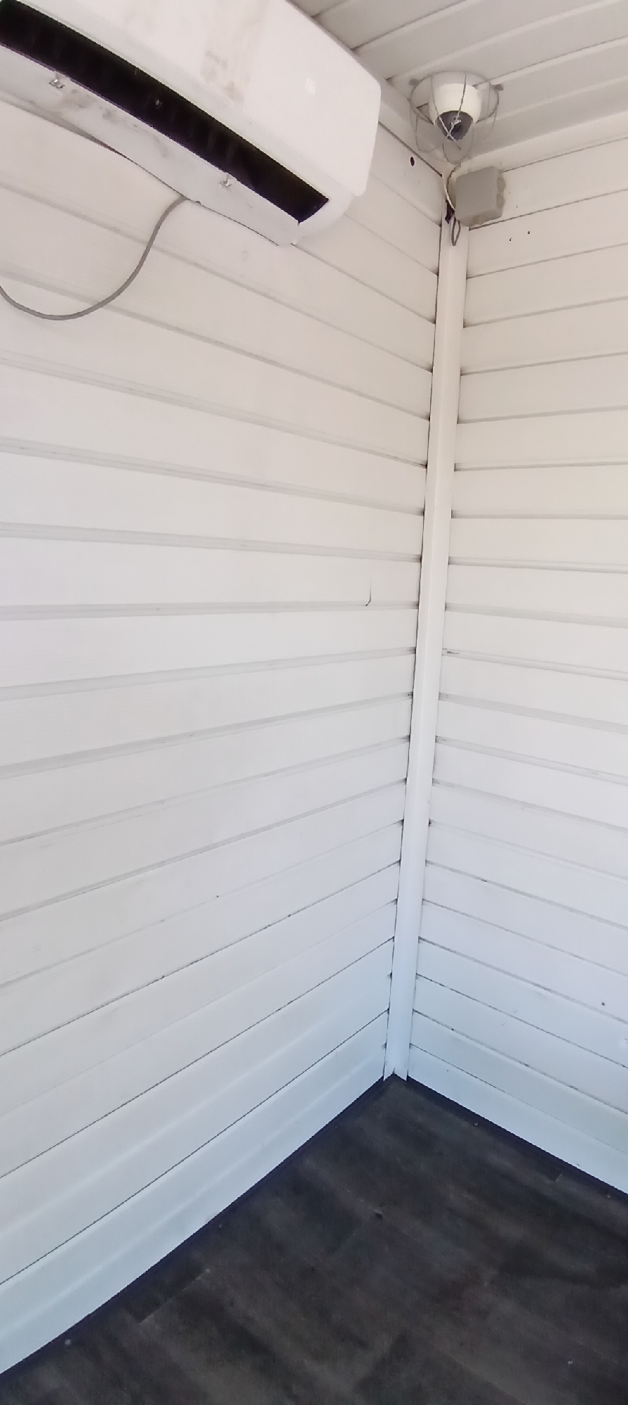 The image shows an interior corner of a room with white paneled walls and a dark wooden floor. An air conditioning unit is mounted high on the left wall. On the right wall, near the ceiling and above a small grey electrical box, a security camera is visible, enclosed within a protective cage. The walls appear to be made of horizontal wooden planks, painted white. The floor is composed of dark, wood-look planks laid horizontally. The scene is well-lit, suggesting an indoor environment. There are no people or animals present in the image. The location is not specified in the image itself.