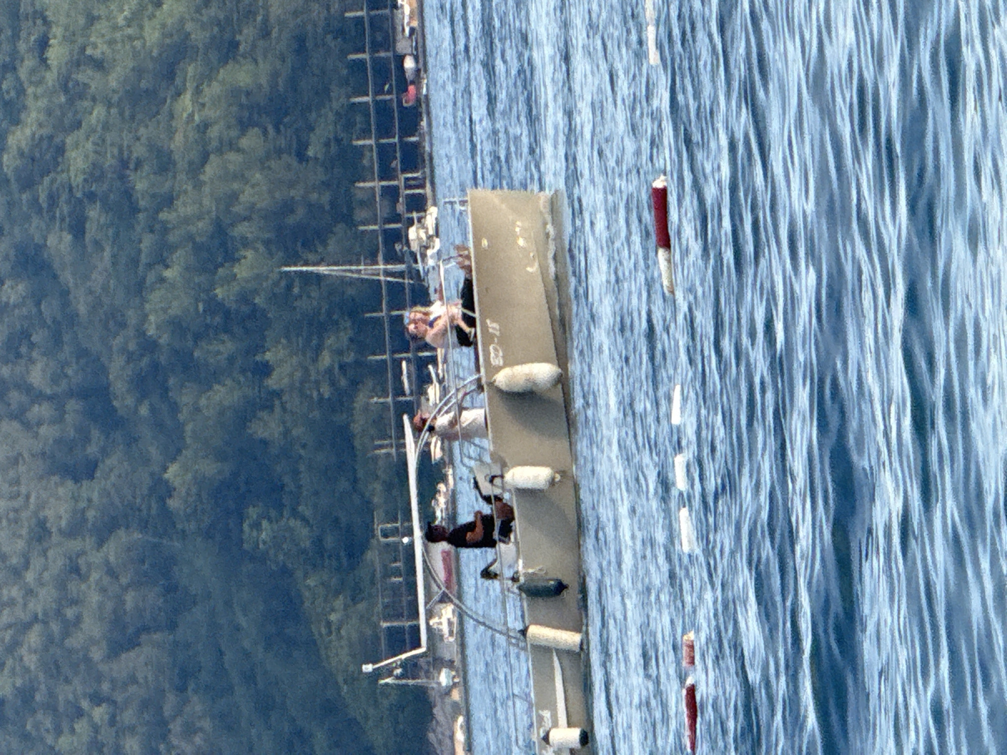 The image shows a view from above of a body of water with a dock or pier extending into it. There are several people visible on the pier, some sitting and some standing. One person appears to be lying down. There are also several buoys in the water, with one red and white buoy prominently featured near the pier. In the background, there is a dense forest covering a hillside. The water is choppy, suggesting it might be a windy day. The overall scene appears to be a recreational area, likely a marina or a lakefront. There is some text visible on the side of the pier, but it is partially obscured and difficult to read clearly, though it appears to be a designation or identification number. The lighting suggests it is daytime, possibly late morning or afternoon, given the bright sunlight on the water and the shadows. The location is identified as Budva, Montenegro.