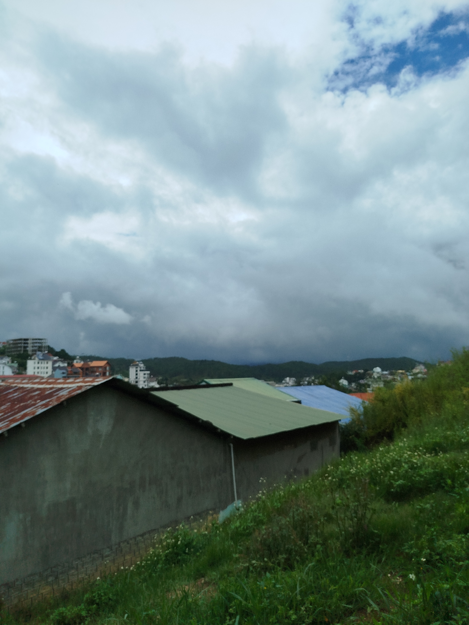 This image captures an expansive, overcast landscape of Da Lat, Vietnam, viewed from an elevated position. The sky dominates the upper two-thirds of the frame, filled with dense, grey clouds that suggest a cool, possibly damp, day. A small, brighter patch of blue sky is visible in the upper right corner, indicating some breaks in the cloud cover.
In the foreground, the scene features parts of two buildings. On the left, the weathered, rusty corrugated metal roof of a structure is visible. Adjacent to it, towards the right, stands a building with a dark green metallic or painted roof, its plain concrete wall blending into the earthy tones.
The mid-ground features a vibrant green hillside covered in wild grasses and small white flowers, sloping downwards into the distance. Beyond this natural expanse, a town or city unfolds across a valley, with numerous buildings nestled amongst forested hills. Visible structures include multi-story white buildings, and others with colorful roofs in shades of blue and orange, reflecting typical residential and urban architecture.
Dark green hills and mountains form the distant background, partially obscured by the extensive cloud cover and atmospheric haze, which lend a serene and somewhat muted atmosphere to the scene. No people are visible, and no specific activities are taking place. The time of day appears to be daytime, but the heavy clouds give it a soft, diffuse light. There is no visible text in the image.