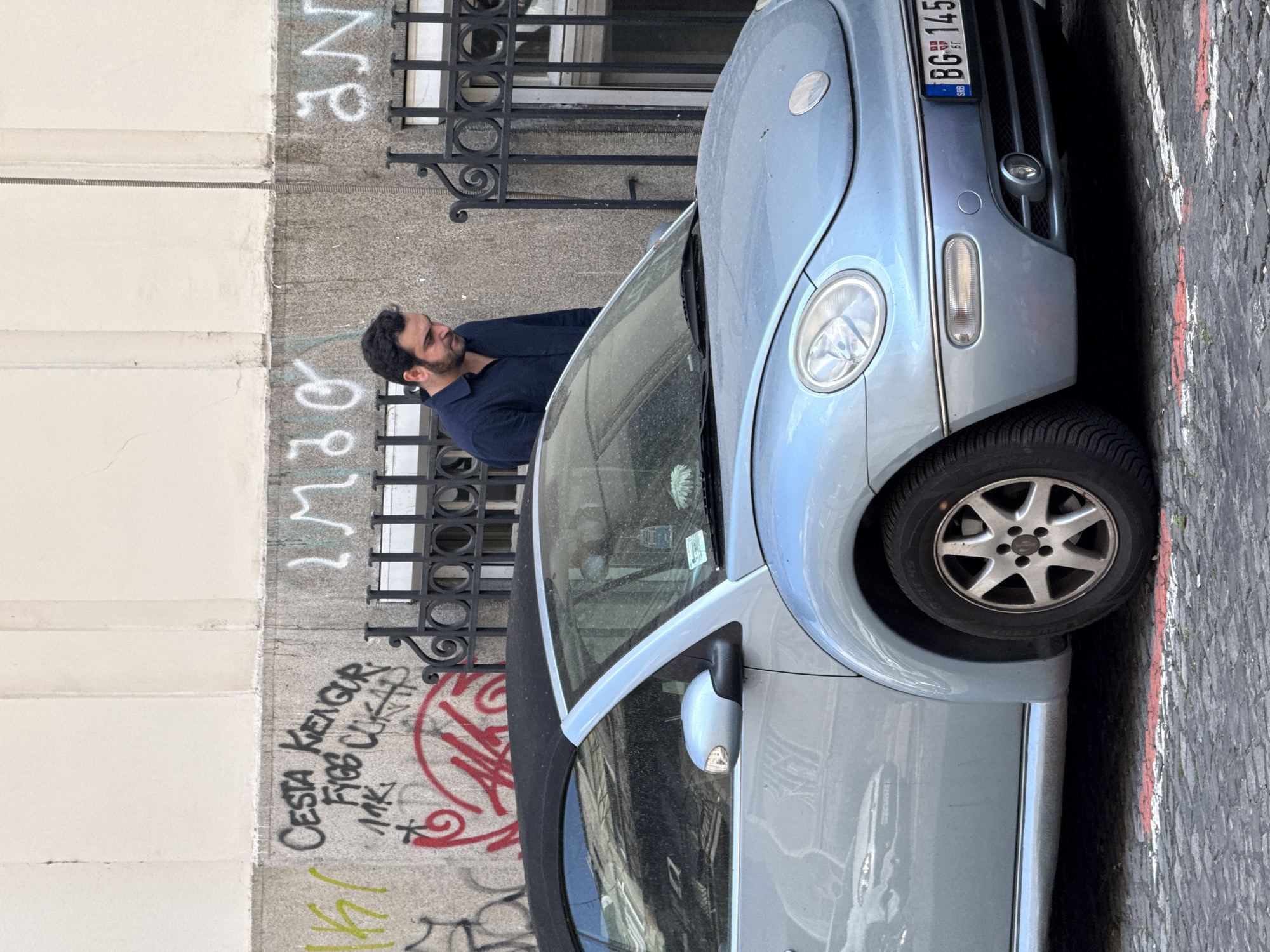 The image captures a daylight street scene in Belgrade, Serbia, featuring a man, a parked car, and a building facade with graffiti.
A man with dark hair and a beard, wearing a dark blue collared shirt, stands in profile on the left side of the frame. He is holding a black smartphone to his right ear, seemingly engaged in a conversation, and leans casually against a black, ornate metal fence or gate.
In front of the man, and occupying much of the right side of the image, is a light blue or silver-grey Volkswagen Beetle convertible. The car is parked parallel to the curb, showing its front passenger side, including the headlight, front grille, part of the wheel with an alloy rim, and a dusty windshield. The convertible top is dark.
The background consists of a building with a light-colored, smooth upper section and a darker, textured lower wall where the man is leaning. This lower wall is covered in various graffiti. The street is paved with grey cobblestones, and a thin red line is visible along the curb next to the car.
Notable details include the car's license plate, which displays "BG 145" and "SRB" with a small Serbian flag, confirming the location. Visible graffiti on the wall includes black text that reads "CESTA KRENUH", "FIBES CUPA", and "MRK", along with other indistinct white and green spray paint markings. The bright, even lighting indicates daytime. The man appears calm and focused on his phone call.