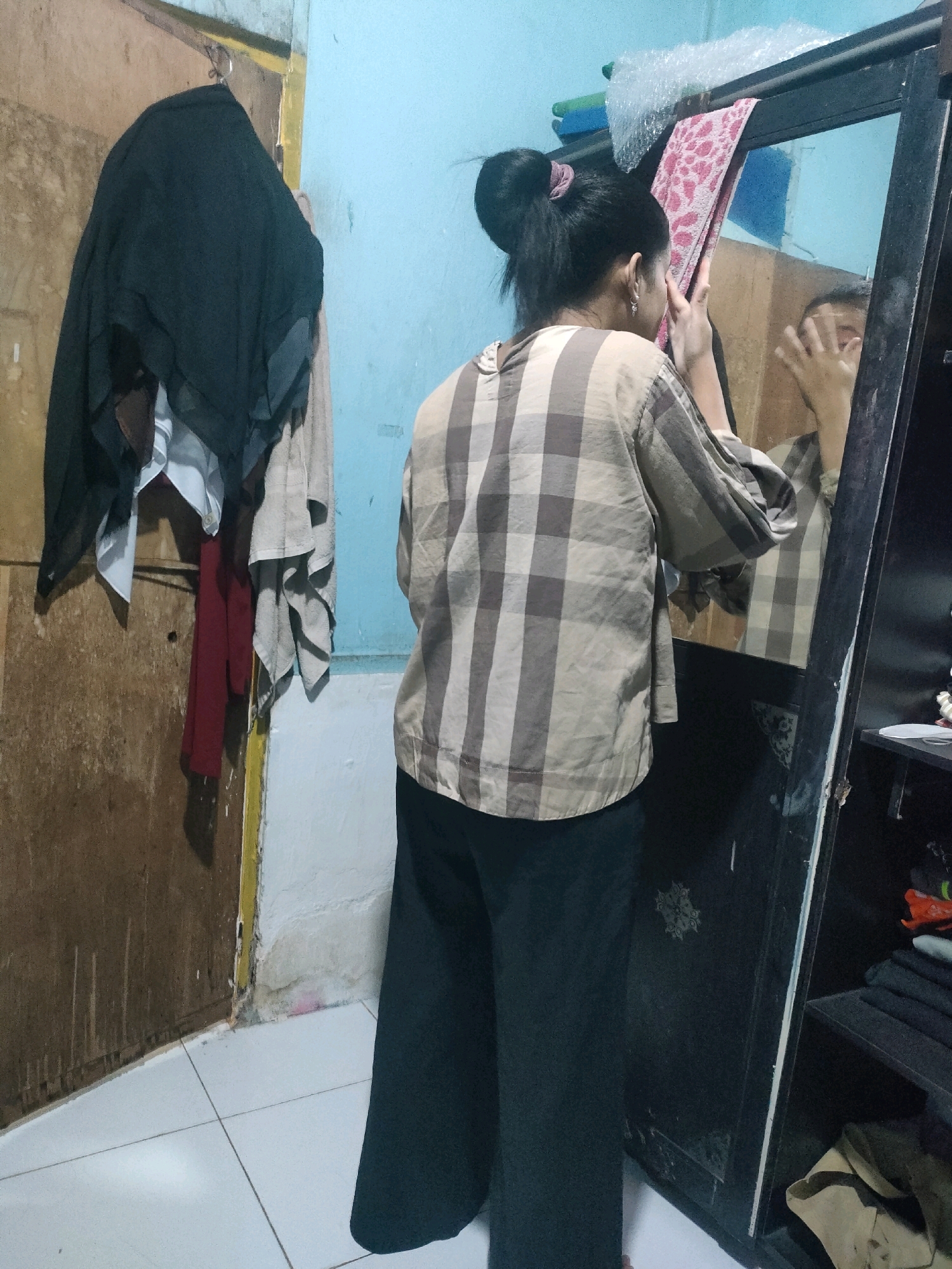 The image depicts an indoor scene, likely a residential room in Pekanbaru, Indonesia. A young woman with her hair tied in a bun is standing in front of a full-length mirror, adjusting a pink patterned towel or cloth on the mirror's surface. She is wearing a light brown and grey checkered long-sleeved shirt and black wide-leg pants. To her left, a wooden door is visible, with several pieces of clothing, including a black hijab and a red garment, hanging from hooks. The room features light blue walls and a tiled floor. The mirror is part of a dark-colored cabinet, which is partially open on the right side, revealing shelves with folded clothing. The overall impression is one of a casual, domestic setting.