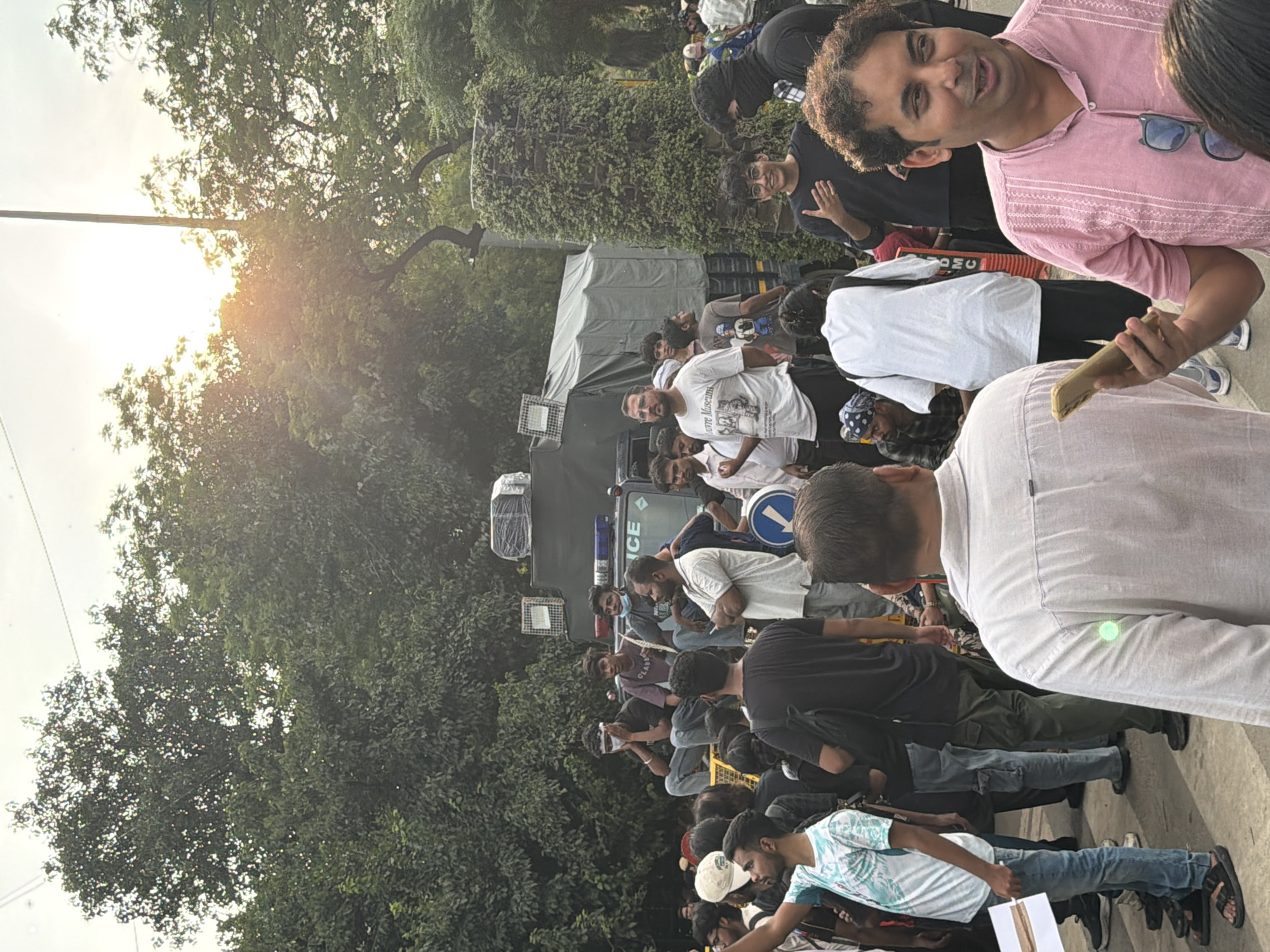 In a city street in New Delhi, India, a diverse group of individuals is gathered. A large military-style truck with a canvas covering is parked in the background, marked with the word "POLICE". Several young men are seated on the truck, some with head coverings or masks. A man in a white t-shirt that reads "Pierre Museum" stands to the left of the truck, looking towards the camera.
In the foreground, a man in a pink shirt holds a mobile phone and gestures with his other hand, appearing to speak. He wears sunglasses clipped to his shirt. Other people are visible in the crowd, some holding signs or phones. The environment includes lush green trees and a cloudy sky, with the sun partially visible through the foliage, suggesting it is late afternoon. The scene appears to be a public gathering or protest.