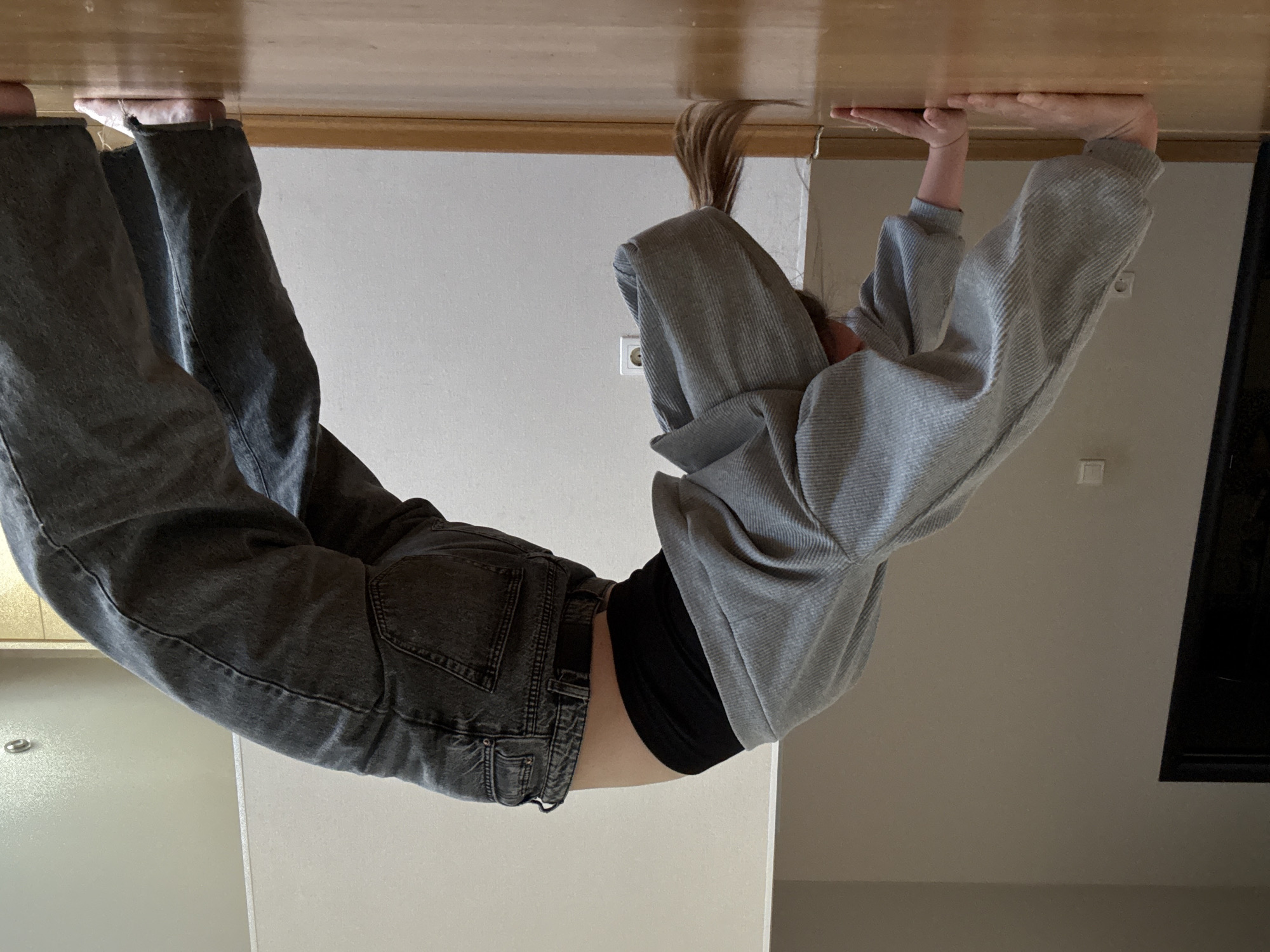 A person is performing a handstand in an interior space. The person is wearing gray jeans, a black top, and a gray sweatshirt. Their hands are pressing against a light-colored wooden surface, which may be a ceiling. The environment includes light-colored walls, an electrical outlet, and a glimpse of a dark object that could be a television or picture frame. It is not possible to determine from the image if the location is in Chaikovsky, Russia.