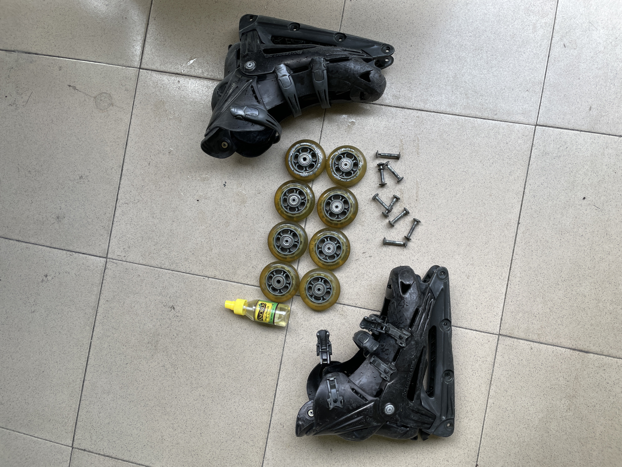 An overhead shot shows a pair of black rollerblades, dissembled, laid out on a light beige tiled floor. The right rollerblade boot is on the left side of the frame, with its laces and straps undone. To the right of the boot, a small yellow bottle of oil with a white label that reads "KNORRIS Extra Virgin Olive Oil" sits near a group of eight tan inline skate wheels, arranged in a two-by-four grid. Below the wheels, a scattering of silver bolts lies on the tiles. The right rollerblade frame is positioned to the right of the wheels, also disassembled. The floor tiles are square and appear to be in an outdoor setting, possibly a patio or courtyard, as there are small specks of dirt visible on some tiles. The lighting suggests it is daytime, with natural light illuminating the scene. The overall impression is that of a DIY rollerblade repair or maintenance session. The scene is located in Ado, Nigeria.
