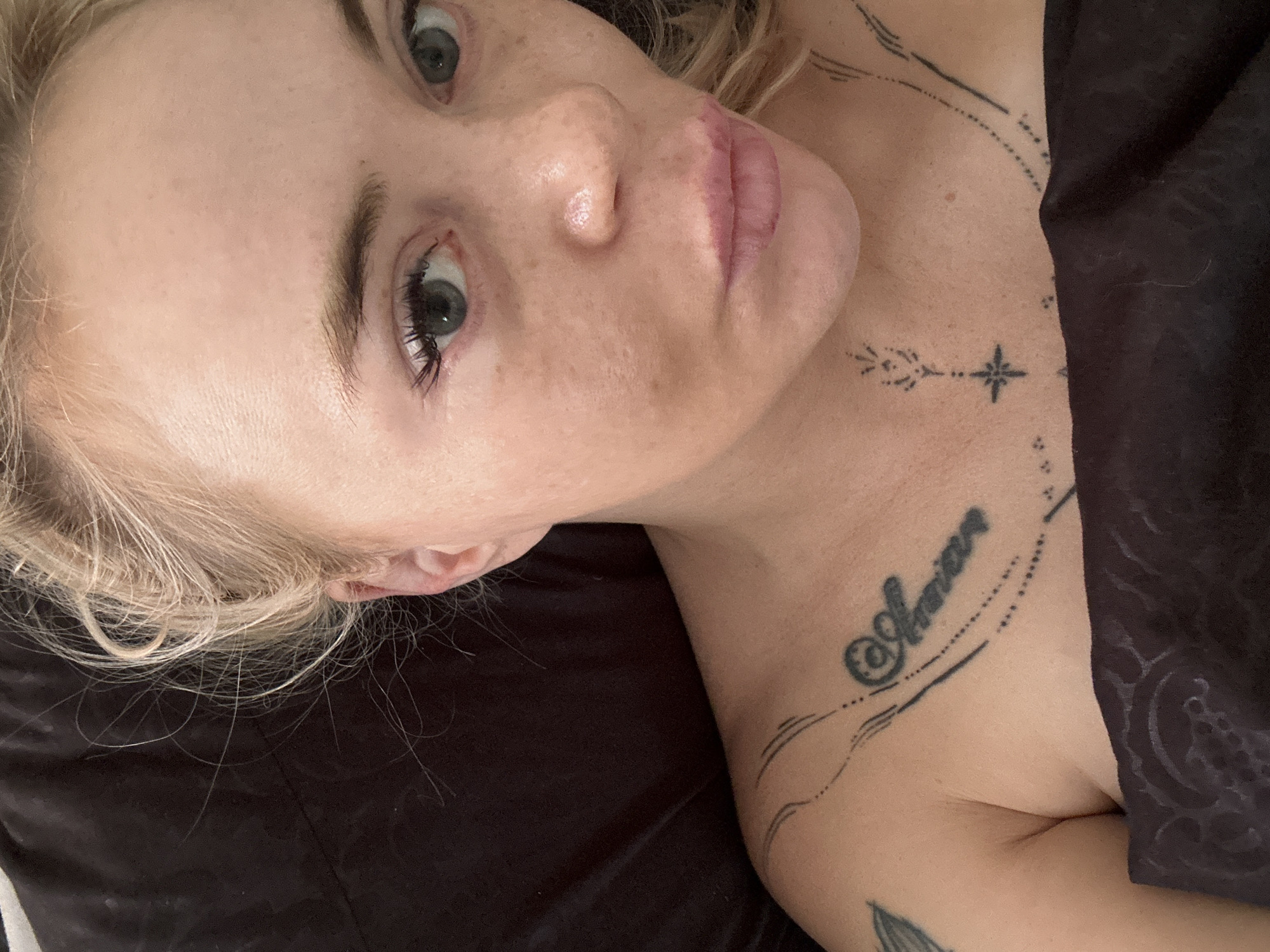 A close-up, overhead shot shows a fair-skinned person with blonde hair lying down and looking towards the camera. The person has freckles on their face, visible eyelashes, and pink lipstick. They are wearing a dark, patterned fabric that covers their chest and shoulders, and a tattoo is visible on their left shoulder. The background is a dark, solid surface. The lighting is soft and appears to be natural.
