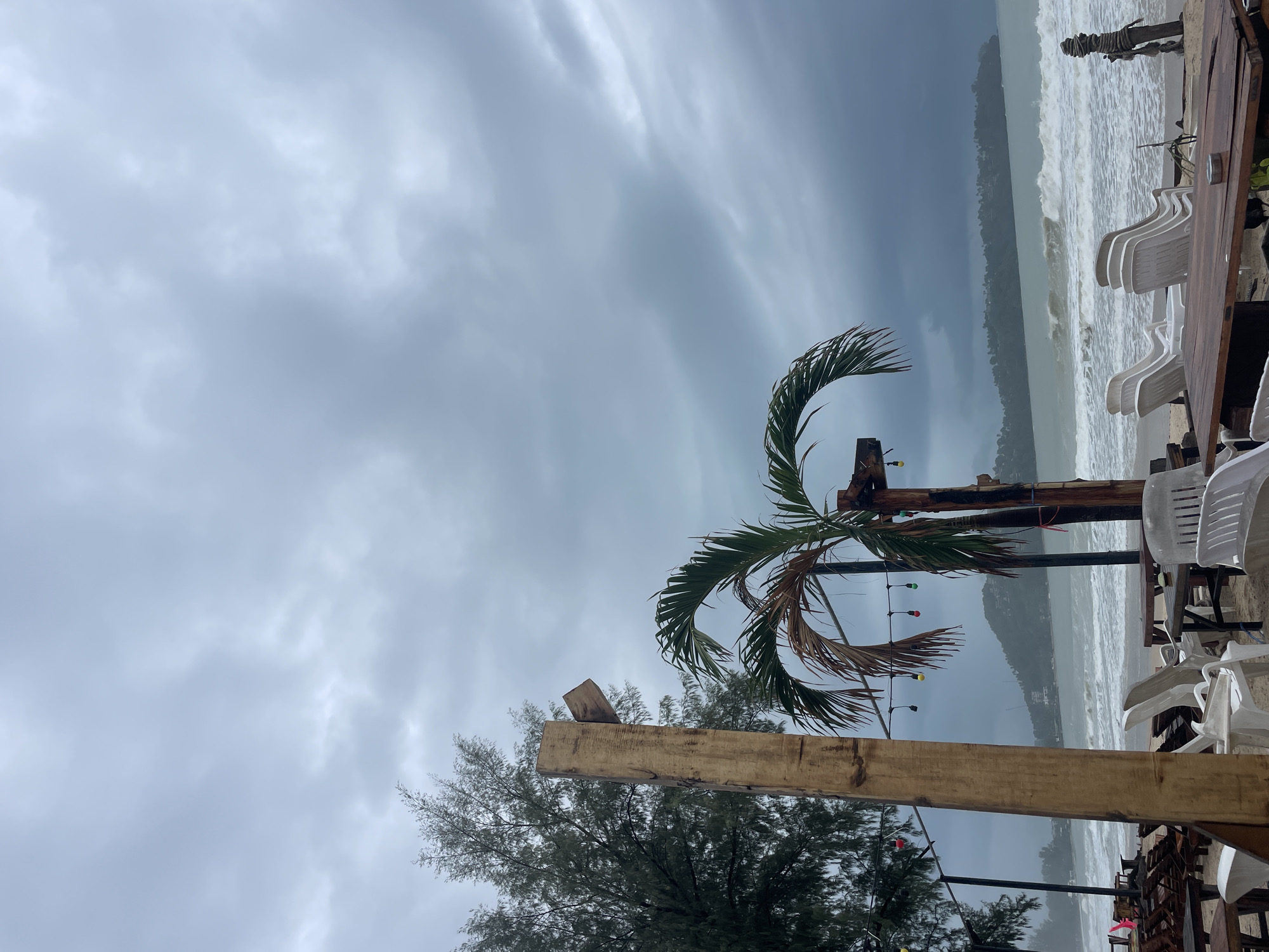 The image captures a beach scene in Choeng Thale, Thailand, under a stormy sky. The sea is rough with large waves. In the foreground, there's a structure made of wooden beams, a small palm tree, and outdoor furniture including white plastic chairs. String lights are wrapped around one of the beams and the palm tree. There are no people visible.