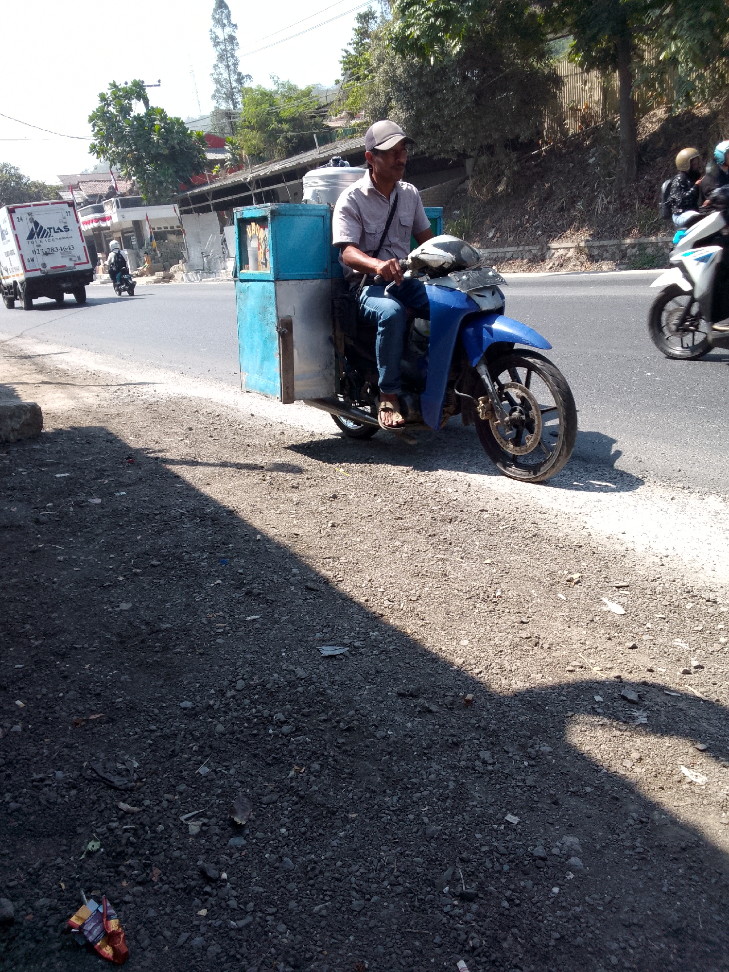 In Gunungmasigit, Indonesia, a man wearing a cap, light-colored short-sleeved shirt, and blue jeans rides a blue motorcycle. The motorcycle is equipped with a large, light blue and silver container attached to the rear, suggesting it is used for vending or transport. The man is looking forward, seemingly in motion. Beside him, on the right side of the frame, another motorcycle carrying two individuals is also moving. In the background, a white delivery truck is visible on the road, and various buildings and trees line the roadside, indicating an urban or semi-urban environment. The foreground is a gravelly roadside with a shadow cast across it.