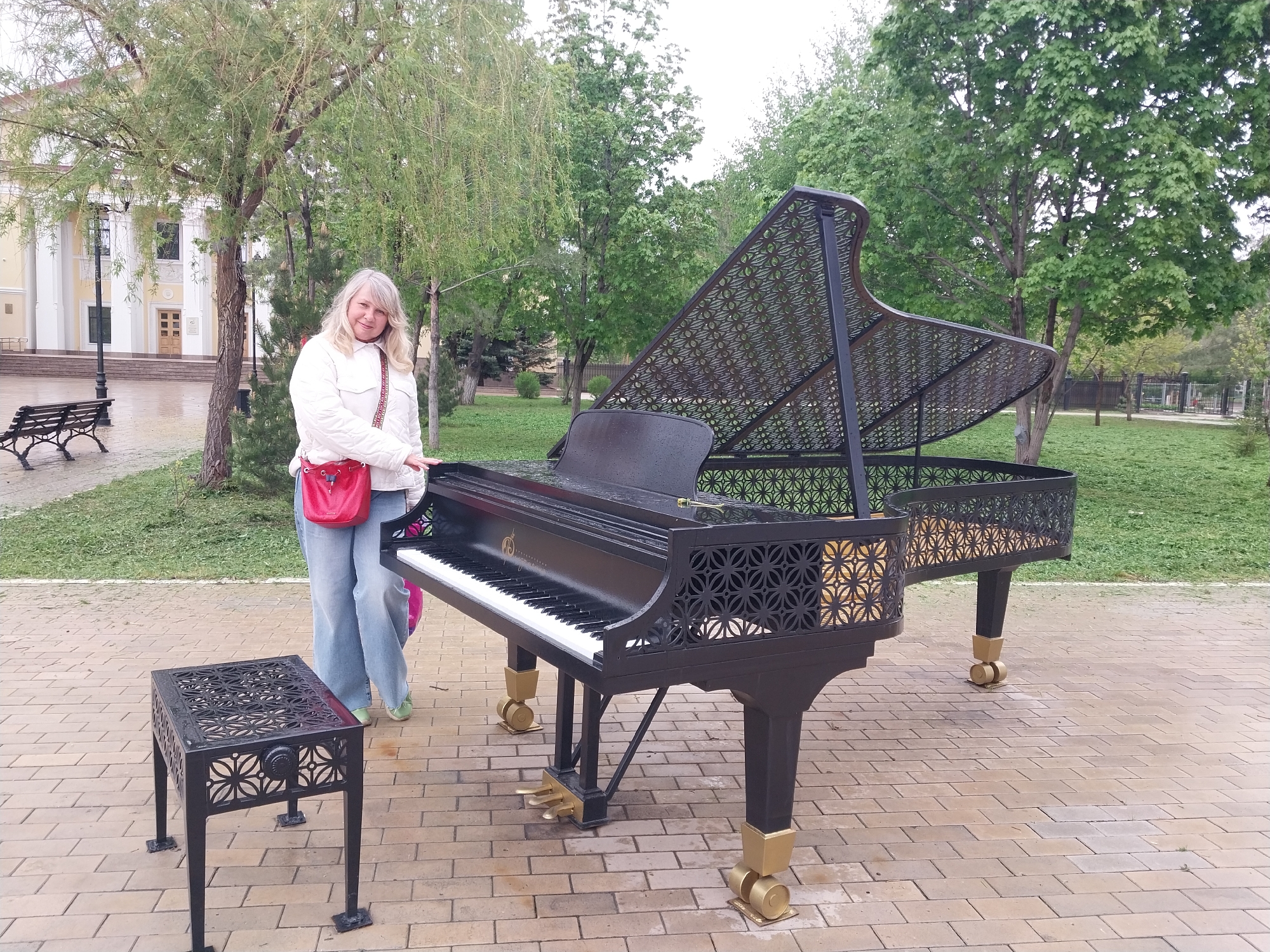 A wide, eye-level shot captures a woman with shoulder-length blonde hair standing to the left of a black wrought-iron piano in a public setting. The woman wears a white jacket, blue jeans, and carries a red purse. The piano, which has an open lid, is detailed with geometric designs and sits on a brick-paved area. To the left of the piano is a matching wrought-iron stool. The background includes green grass, trees, and a building with white columns in the distance. Based on available knowledge, this scene is set in the city of Orenburg, Russia.