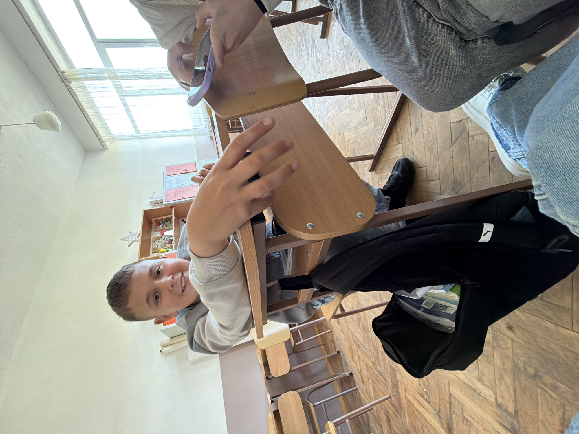 The image captures a classroom scene, likely in Zmeitsa, Bulgaria. A young boy is seated at a wooden school desk, smiling directly at the camera. He wears a grey and white hooded sweatshirt. His desk is one of a row of similar desks on a wooden floor with a herringbone pattern. Another student, wearing a light-colored top, is partially visible at another desk, using a purple tool. A black Puma backpack is hanging on the back of the boy's chair, with books visible inside. A window with multiple panes is visible in the background, providing natural light. There's a shelf with decorative items and possibly books on the wall.