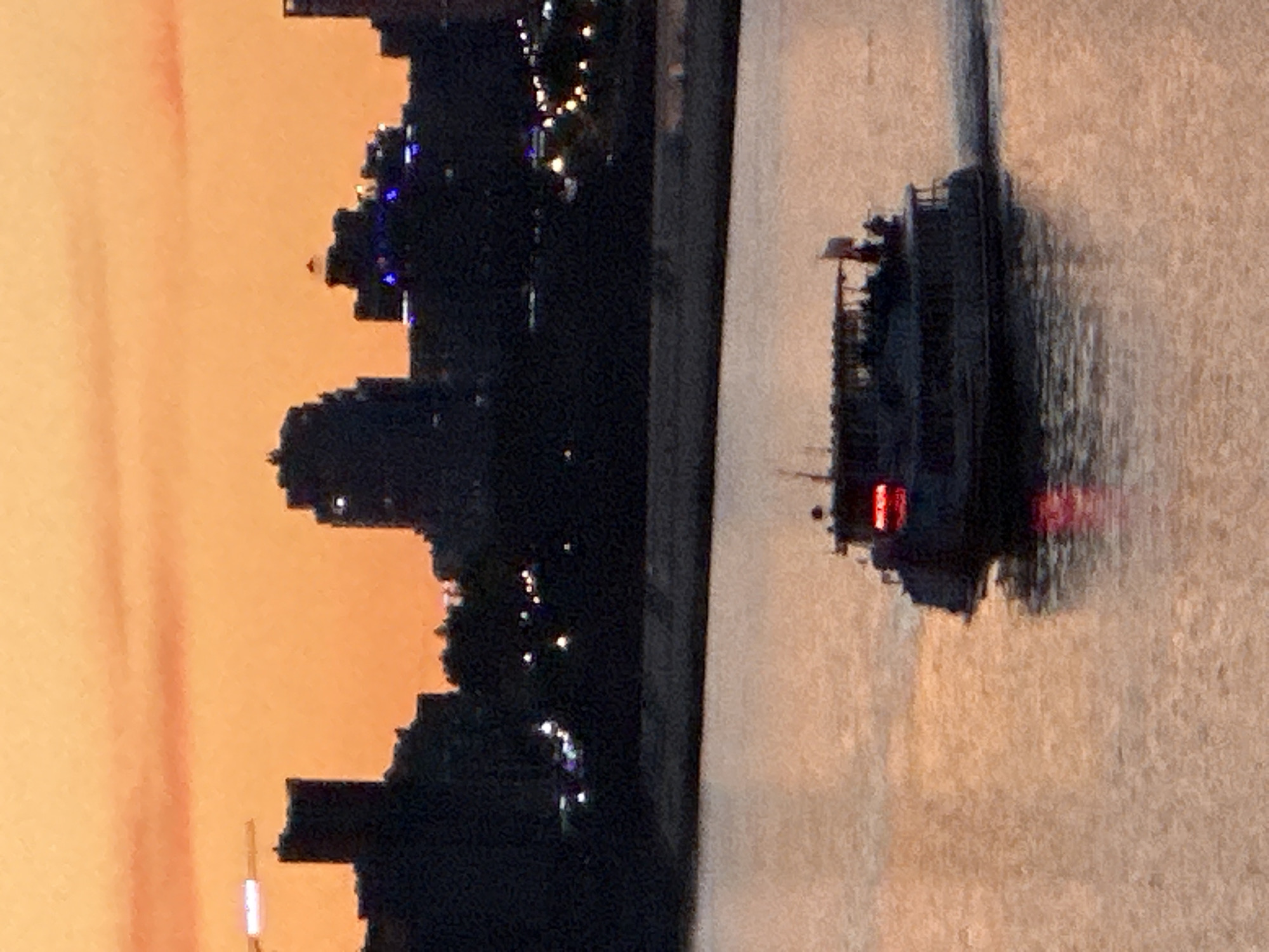The image captures a dusky scene over the Amur River in Khabarovsk, Russia. Silhouetted against an orange sky, the city skyline is visible, featuring several tall buildings and structures. In the foreground, a boat, possibly a ferry or tour boat, cruises on the water, leaving a wake. The boat has illuminated windows and red lights visible on its structure. The water reflects the warm hues of the sky, creating a tranquil atmosphere.