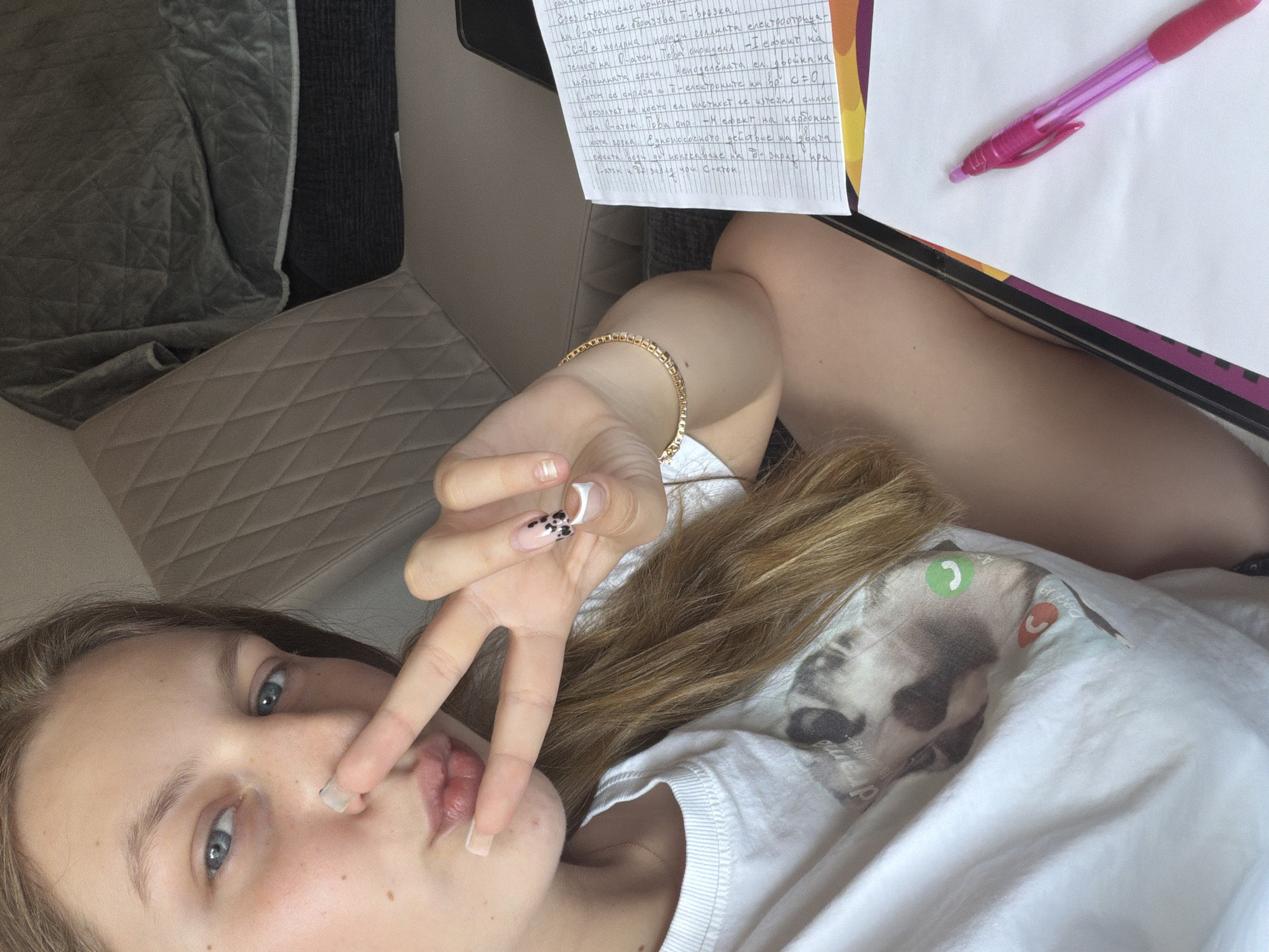 This is a close-up, overhead shot of a young person lying down, likely in a home setting. The person has light brown hair and blue eyes, and is wearing a white t-shirt with a graphic of a cat's face. They are making a peace sign with their right hand, which has a gold bracelet on the wrist and decorative nail art on the fingers. The person is touching their nose with their index finger, and their lips are slightly parted, giving them a playful or relaxed expression.
In the background, there are some papers with handwritten text in what appears to be Bulgarian, suggesting the person might be studying. A pink pen rests on top of the papers. The scene is set on a textured surface, possibly a couch or bed, with a quilted grey blanket visible to the left. The lighting suggests it's daytime. The overall mood is casual and relaxed, with a hint of study or creative work. The visible text appears to be related to physics or chemistry concepts, with terms like "atom," "electron," and "bond."