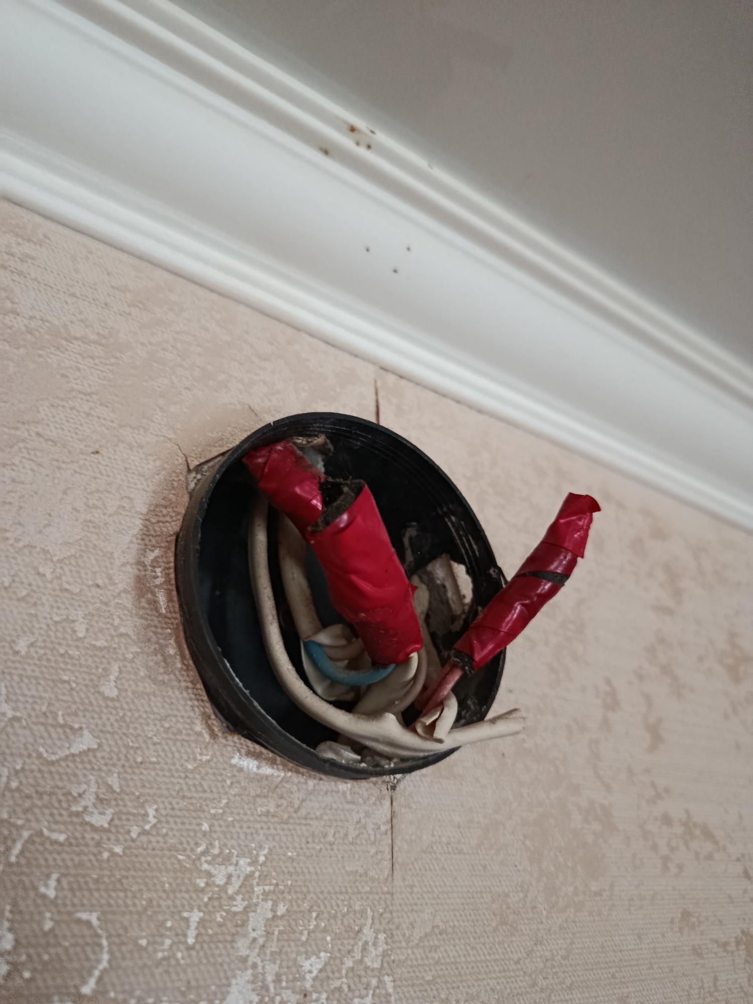 The image shows a close-up of electrical wires protruding from a circular black junction box mounted on a textured, light-colored wall. Two sets of wires are visible, each individually wrapped in red electrical tape. One set is more extensively wrapped and appears to be frayed at the end. The other set is also wrapped in red tape with black tape visible in places. The junction box is partially obscured by the wires and appears to be damaged or worn. The wall has a subtle pattern and some peeling or discoloration. Above the junction box, a white crown molding with a decorative profile is visible, with several small dark specks on its surface. The overall impression is one of unfinished or damaged electrical work. The location is specified as Petropavl, Kazakhstan.