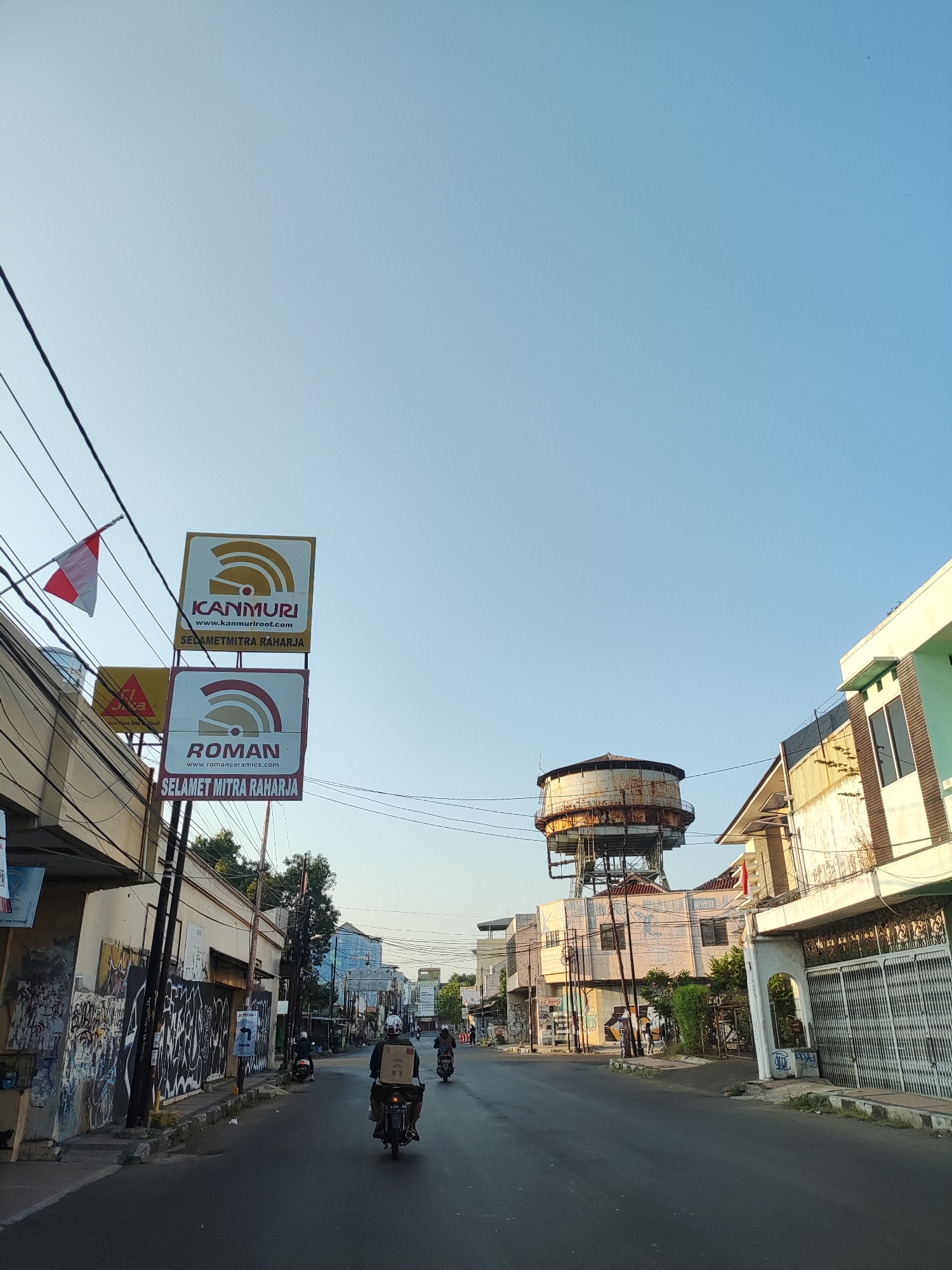 The image depicts a street scene in Cirebon, Indonesia, likely during the late afternoon or early morning given the clear blue sky and soft lighting. On the left side of the street, several signs are prominently displayed, including one for "KANMURI" and another for "ROMAN" with accompanying URLs and the phrase "SELAMET MITRA RAHARJA". A small Indonesian flag flies from a pole above these signs. The buildings lining the street appear to be a mix of commercial establishments, some with graffiti on their lower sections. Further down the street, a large, rusty water tower stands on a metal support structure. Several motorcycles are visible, with some riders carrying packages, suggesting delivery or transportation activity. The road surface is asphalt, and there are power lines running overhead. The overall impression is of a moderately busy urban street with a slightly weathered aesthetic.