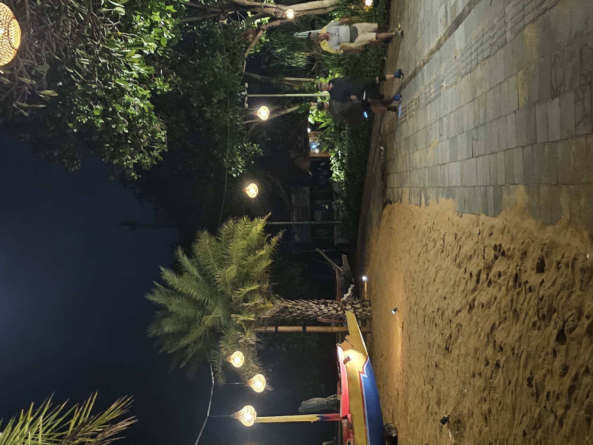 The image depicts a scene in Denpasar, Indonesia, at night. A pathway of light-colored rectangular pavers is partially visible, leading towards an area with trees and shrubbery illuminated by hanging decorative lights. Two individuals are standing or walking on the path. Sandy ground occupies a large portion of the foreground, where a colorful boat sits on the sand. A palm tree stands between the boat and the lights hanging above the shrubbery. Additional palm fronds are visible in the lower left corner. The scene is lit by the decorative lights and potentially other artificial lighting sources.