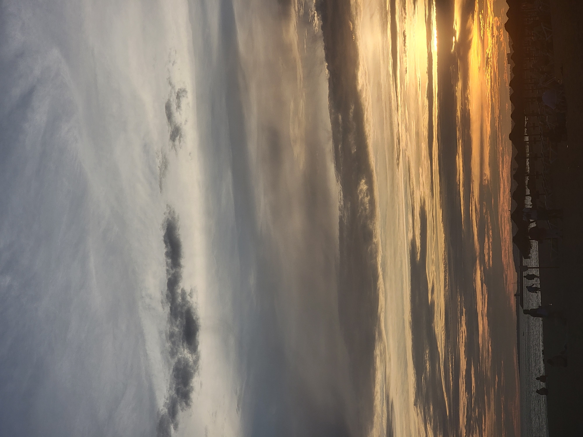 A sunset scene unfolds over the beach and ocean in Quarteira, Portugal. The sky displays a dramatic gradient of colors, transitioning from a pale grey at the top to a vibrant orange and yellow near the horizon. Wispy clouds are scattered throughout, with darker, more defined clouds forming horizontal bands across the middle of the frame.
In the foreground, silhouetted figures are visible on the sandy beach. Several people are walking and standing, some appearing to interact with each other. Beach umbrellas with thatched roofs are set up in rows, with lounge chairs beneath some of them. The ocean stretches out to the horizon, with a faint indication of distant land or structures. The overall atmosphere suggests the end of a day at the beach, with people enjoying the last light.