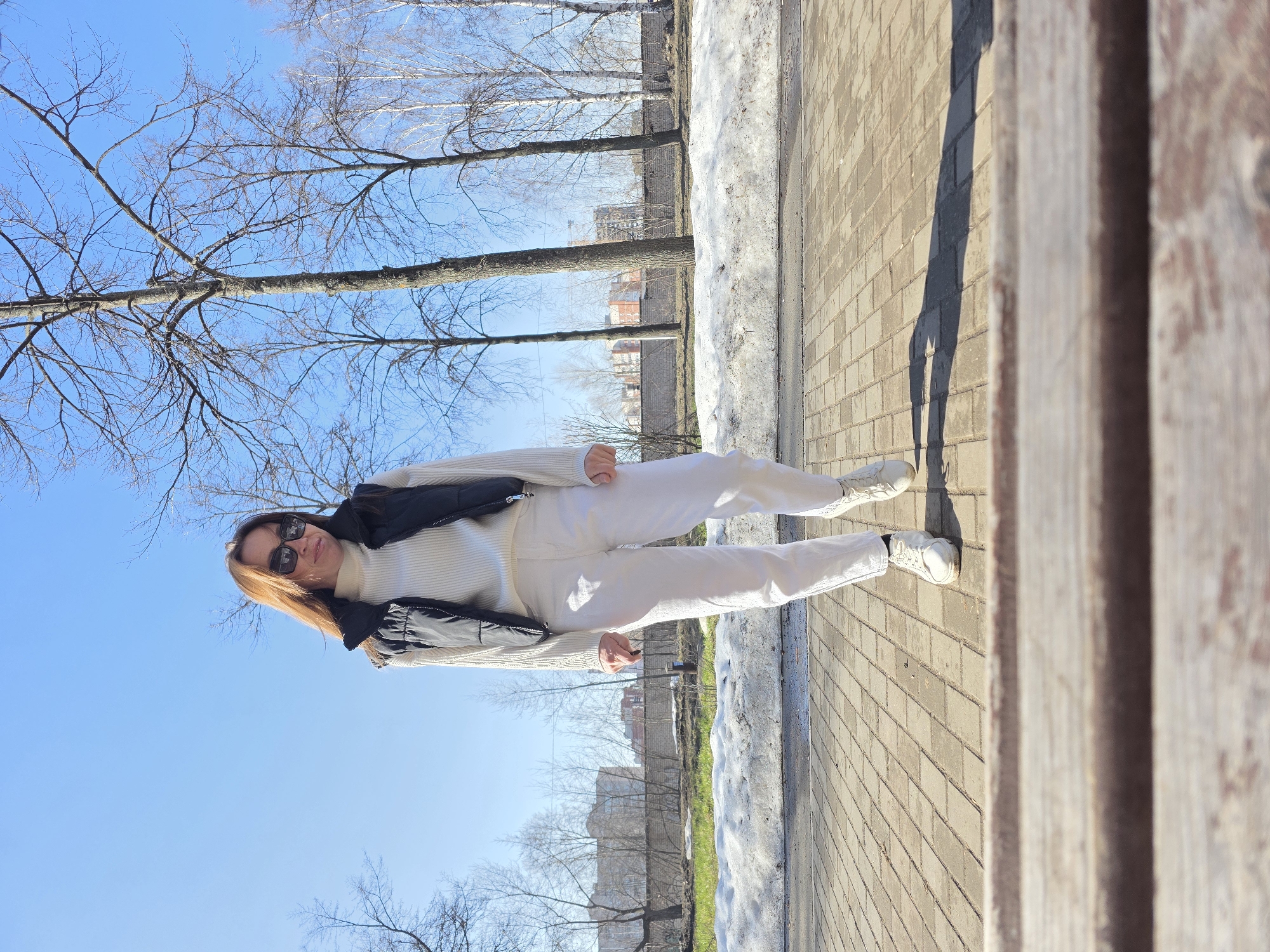 A woman in a white sweater and pants, wearing sunglasses and a black vest, stands on a brick sidewalk. Snow patches are visible near the sidewalk. Bare trees and buildings are in the background. The sky is blue. The location is in the city of Chaikovsky, Russia.