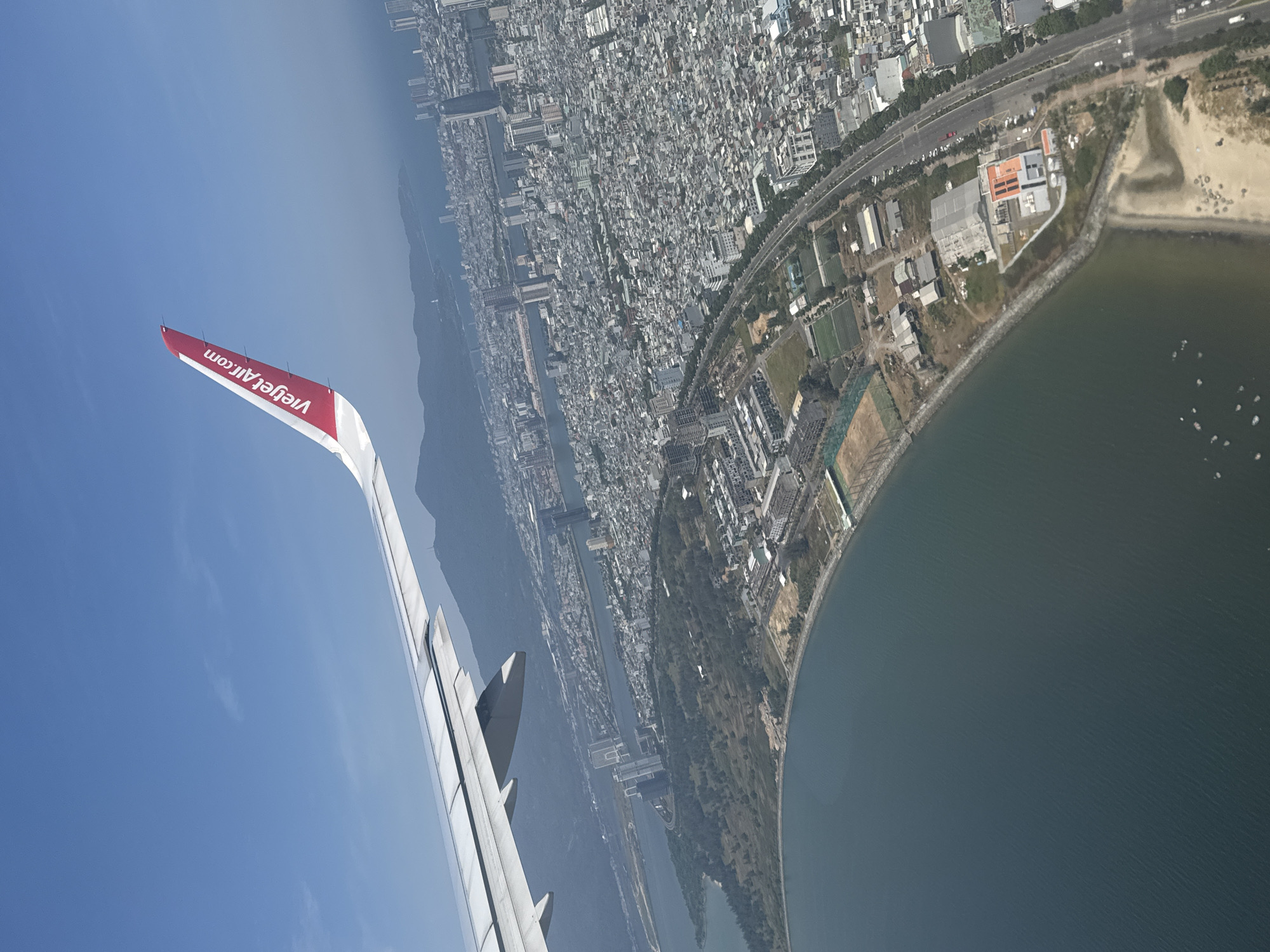 The image shows an aerial view from an airplane window, displaying part of a Vietjet Air plane wing in the foreground and a coastal urban landscape in the background. The wing, with its red tip marked "VietjetAir.com", cuts across a blue sky. Below, a dense cityscape stretches inland from the coast, featuring a mix of buildings, roads, and green spaces. The coastline curves along a body of water, where small boats are visible. This is the city of Đà Nẵng, Vietnam.