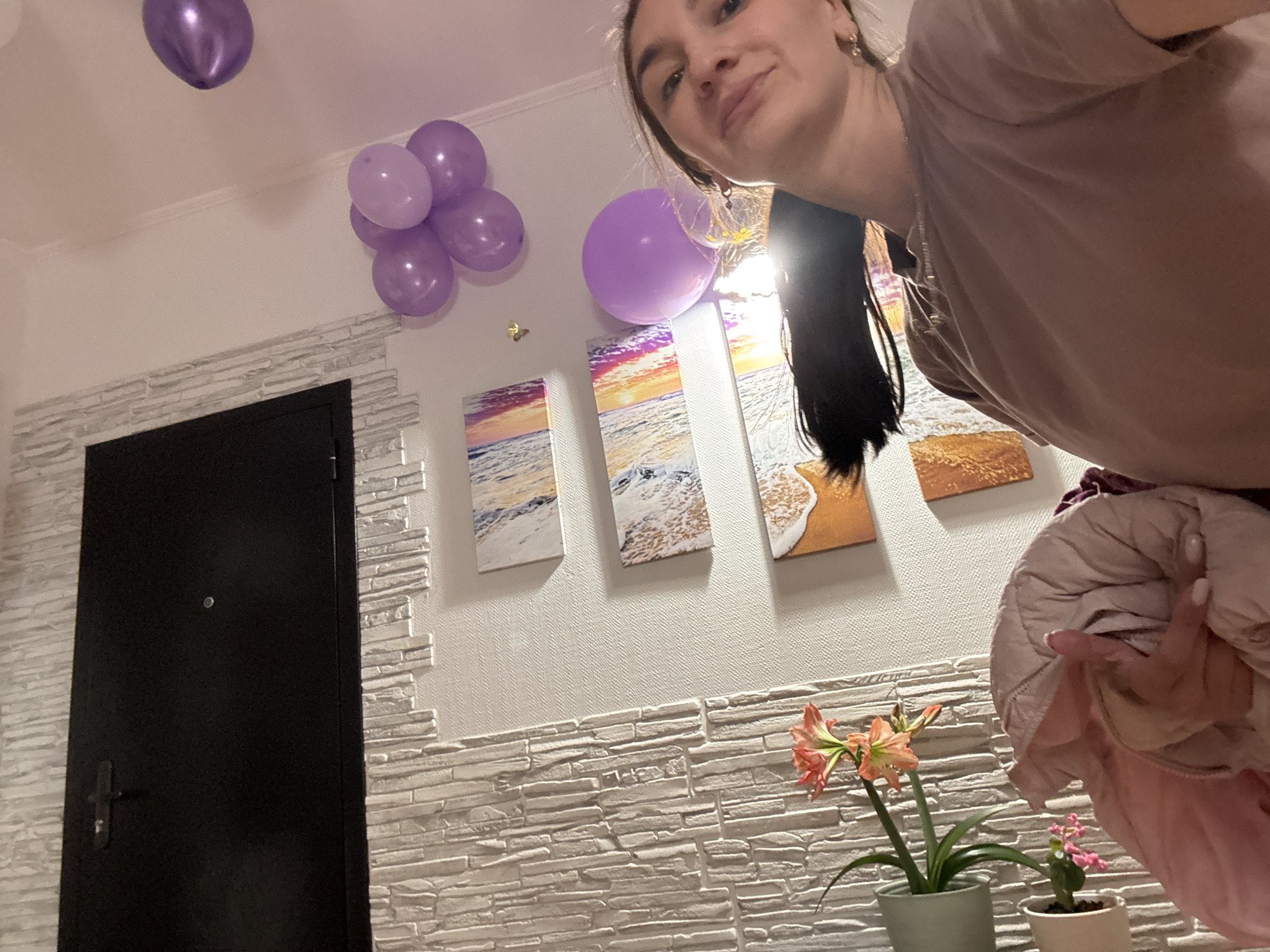 A medium shot captures a young woman indoors with celebratory balloons, art, and potted plants. She's smiling slightly, looking towards the camera, with her dark hair draped over her shoulder. She wears a light brown shirt.
Above the woman and to the left, floating near the ceiling, is a cluster of purple balloons. Below the balloons, mounted on a wall with textured brick-like siding, are three landscape art pieces featuring beach scenes. A black door with a silver handle is positioned on the left side of the frame.
In the foreground, there are potted plants, one with pink flowers and another with a flowering plant. The setting appears to be a residential space, potentially an apartment. The photo does not confirm the location as Sochi, Russia.