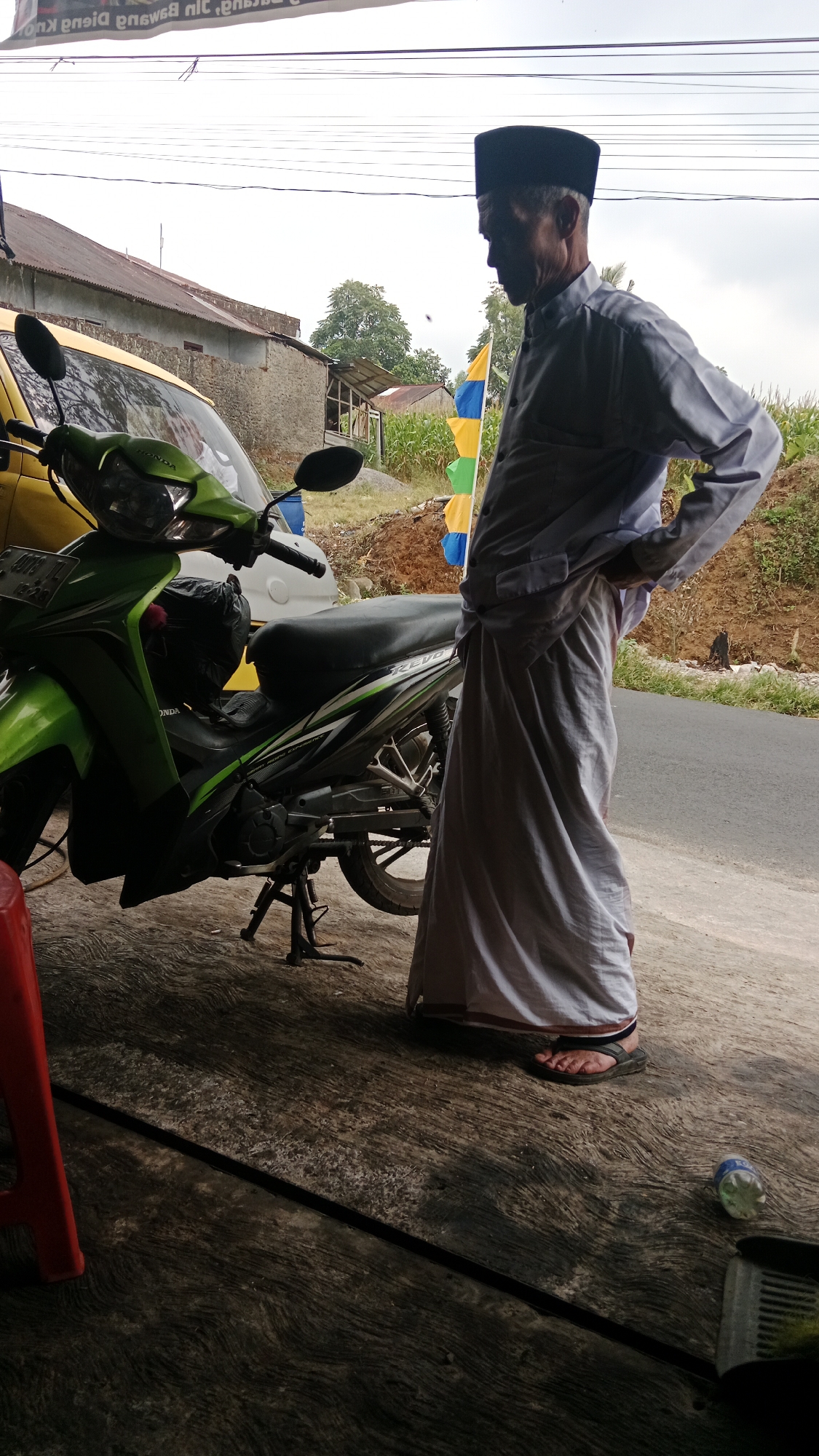 In Candigugur, Indonesia, an elderly man wearing a black peci cap and a white long-sleeved shirt and sarong stands next to a green and black motorcycle. He is facing to the right, with his hands on his hips. The motorcycle is parked on a textured concrete surface, with its kickstand down. In the background, a yellow vehicle is partially visible, along with a stone building and a patch of dirt. There are also colorful flags on poles and some greenery, including corn stalks. The sky is overcast.