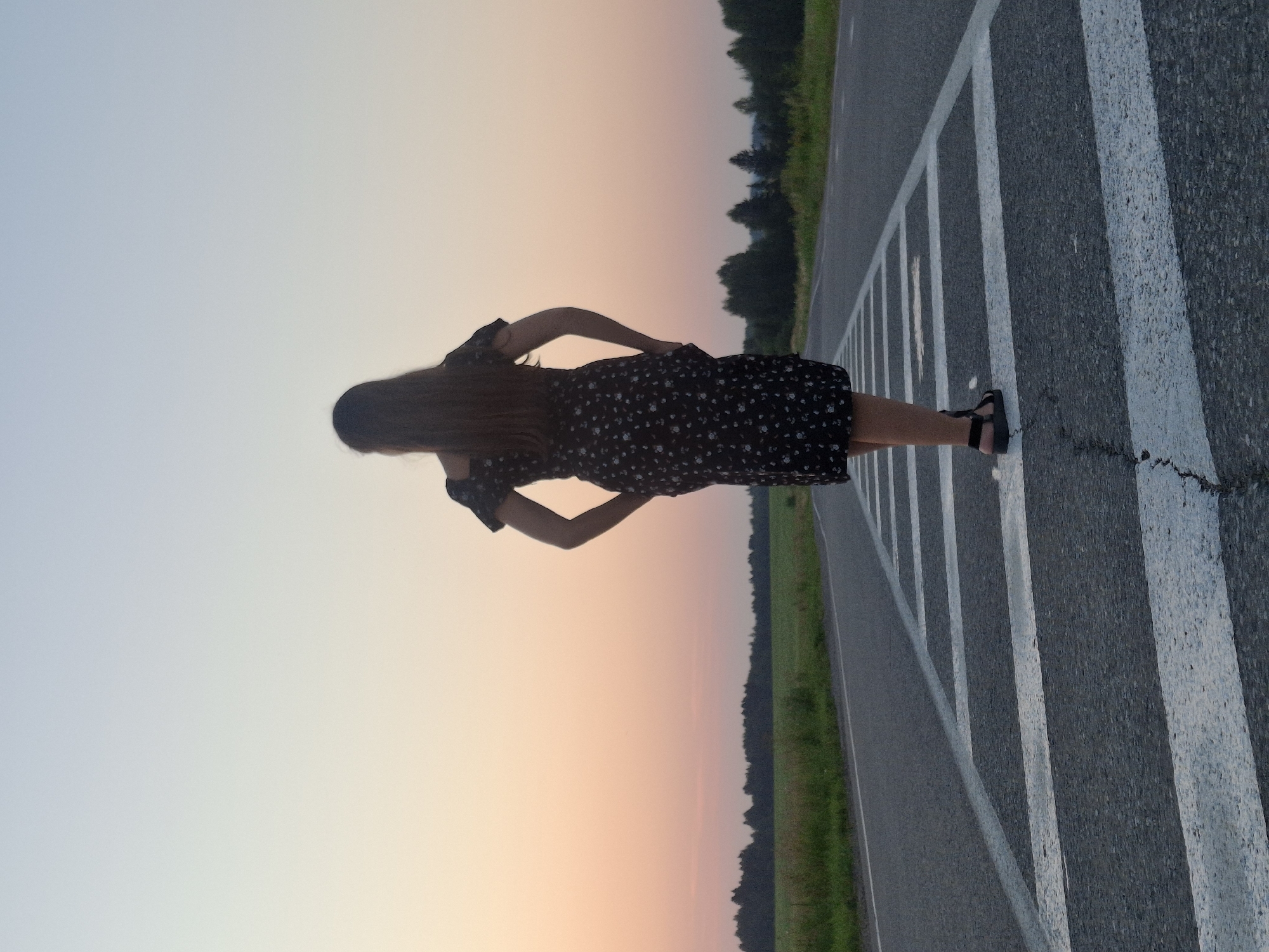 A young woman with long, dark hair stands on a paved road with white crosswalk markings, facing away from the camera. She is wearing a dark, floral-patterned dress and black sandals. Her hands are in her pockets. The road stretches out before her, with a line of trees in the distance. The sky above is a gradient of pale blue and soft orange, suggesting either sunrise or sunset. The scene appears to be in a rural or semi-rural setting, possibly on the outskirts of Perevoznoe, Russia, given the natural landscape and open road. The lighting indicates it is either early morning or late evening.