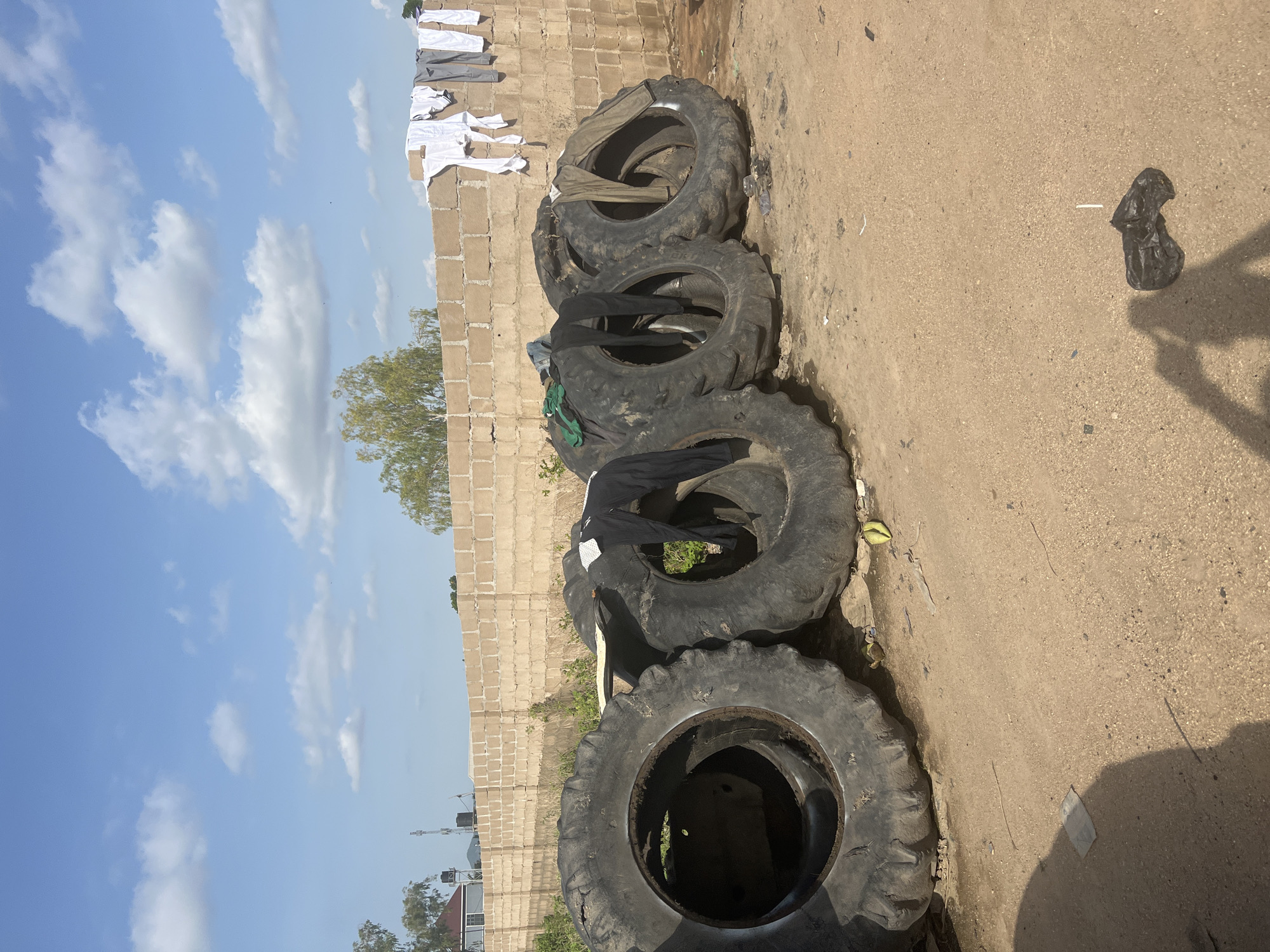 In Jalingo, Nigeria, a line of five large, worn tires is positioned on a dusty, dirt ground in front of a low brick wall. Laundry, including white shirts and darker trousers, is draped over some of the tires and also hanging on a clothesline attached to the top of the wall. The wall is made of beige, rectangular bricks and extends horizontally across the image. Behind the wall, trees are visible, and beyond them, a light blue sky with scattered white clouds provides the backdrop. The overall scene suggests an outdoor area, possibly a residential or communal space, where laundry is being air-dried.