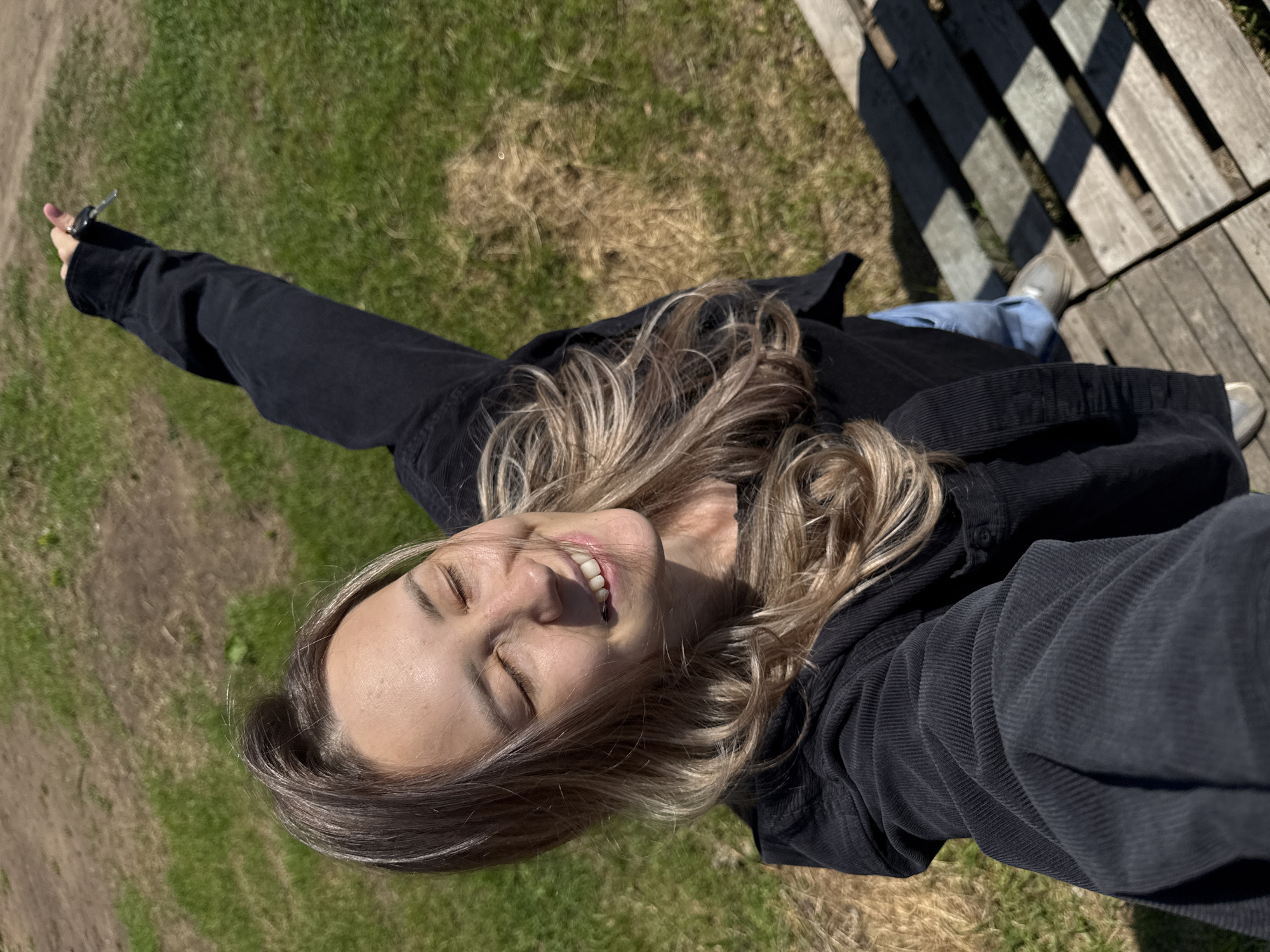 The image depicts a close-up, upward-facing selfie of a young woman outdoors, taken in Chaikovsky, Russia. The woman is smiling broadly with her eyes closed, conveying a sense of happiness and relaxation under bright sunshine. Her long, wavy hair, featuring blonde highlights and light brown roots, frames her face and shoulders. She is wearing a dark, likely black or very dark grey, corduroy jacket.
Her right arm is extended to the left side of the frame, holding a set of keys that include a silver metallic key and a dark fob or ring. Her left leg, clad in light blue jeans and a light-colored shoe, is visible in the lower right portion of the frame, near a wooden structure. This structure, appearing to be a wooden pallet or pathway, is on the right side of the background and casts strong, distinct shadows, indicative of direct sunlight around midday or early afternoon. The ground in the background is covered with a mix of vibrant green grass and patches of dry, brown grass. No text is visible in the image. The overall impression is one of enjoying a sunny moment outdoors.