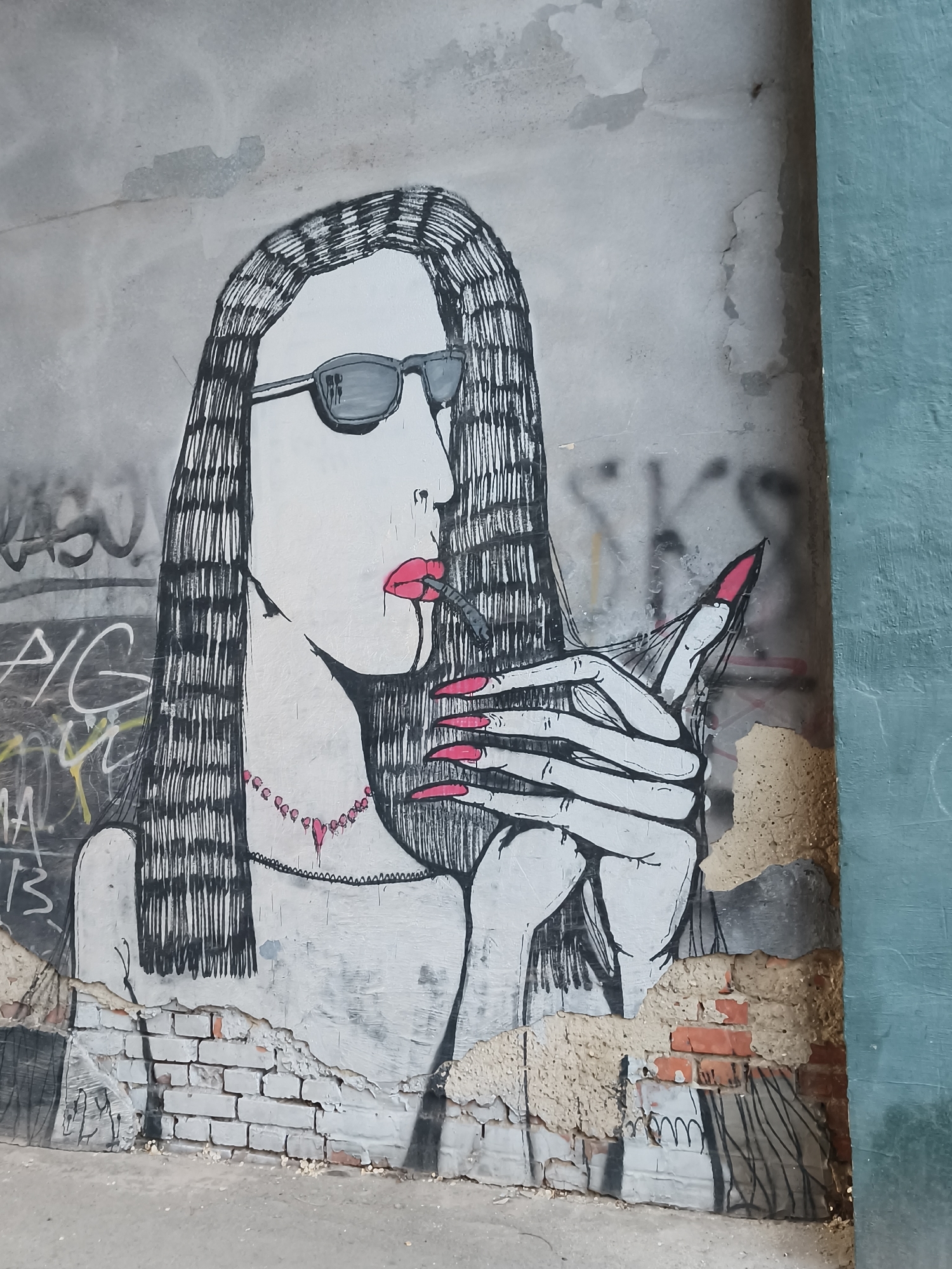 The image shows a black and white graffiti art mural of a woman with long hair, sunglasses, and bright red lipstick and nails. She's holding a thin object between her fingers as if she's smoking a cigarette. The mural is painted on a weathered wall with visible brick underneath in places. The painting is partially damaged, especially at the bottom, revealing the brick underneath. A blue-painted wall is visible on the right edge of the image. Other graffiti is partially visible in the background. Based on available information, this is located in Kryvyi Rih, Ukraine.
