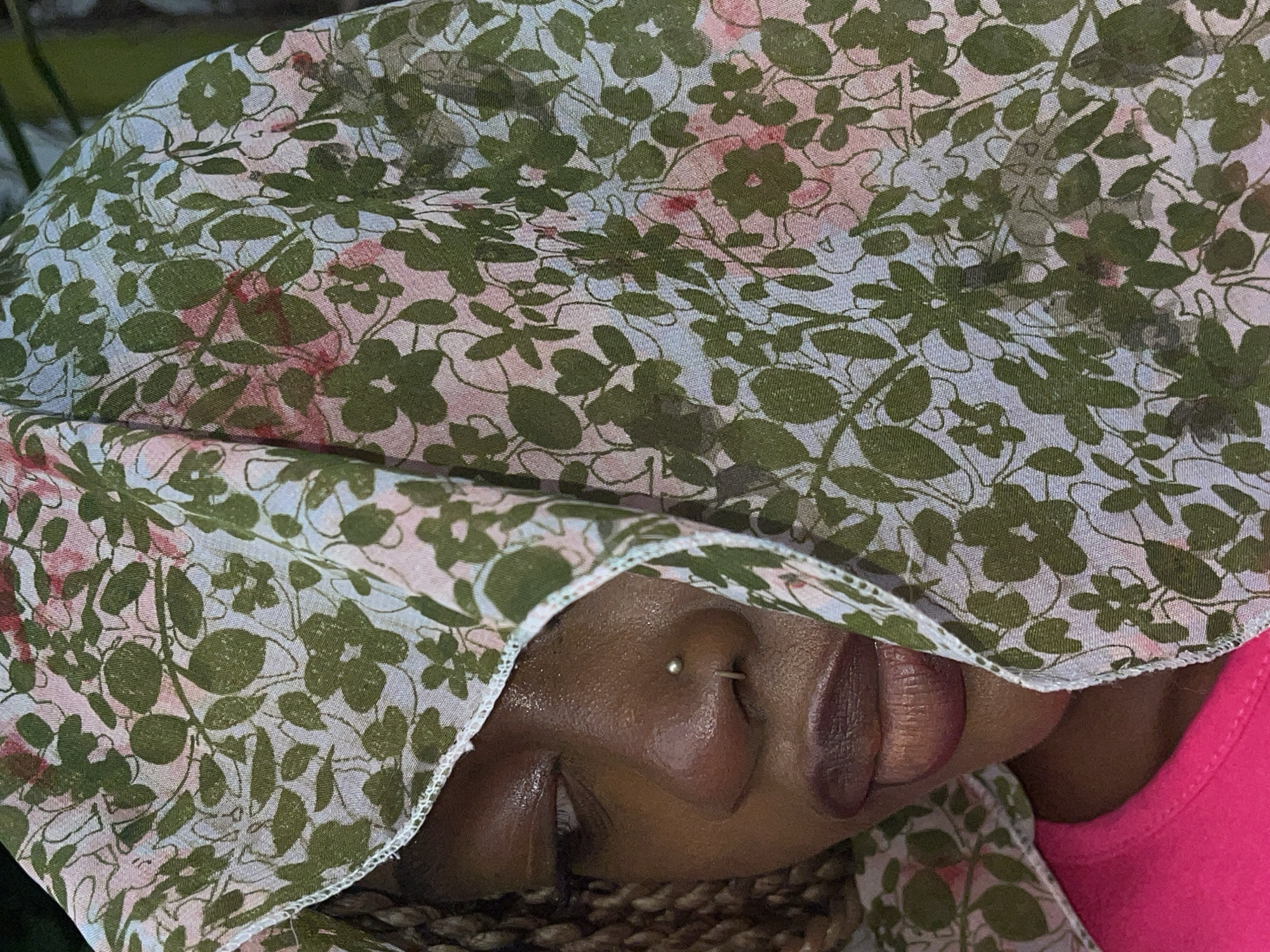 A close-up, angled shot captures the lower portion of a person's face, partially obscured by a floral-patterned head covering. The person has dark skin, and a small, silver stud pierces their left nostril. The lips are slightly parted, revealing some internal texture. Below the chin area, braided hair or locs are visible. A vibrant pink garment, likely a top, is seen on the right side of the frame. The head covering features a design of olive green leaves and subtle pink flowers on a light background, appearing to be a thin, slightly sheer fabric. The lighting suggests it is daytime. The image provides no specific cues regarding activity or emotion, focusing instead on the textures and patterns of the scarf and the visible facial features. The provided location context is Ado, Nigeria.
