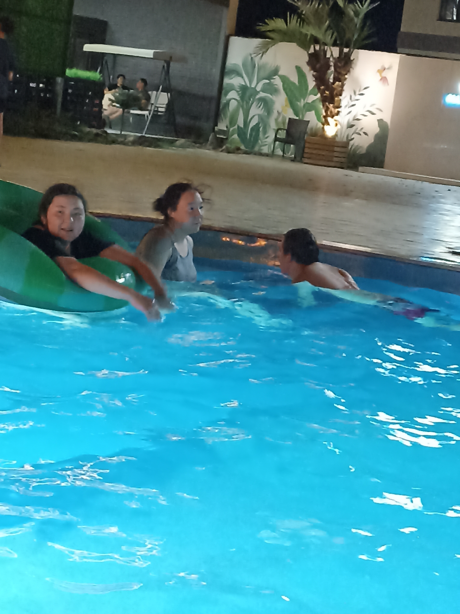 In a nighttime outdoor scene, three individuals are present in a brightly lit blue swimming pool, possibly at a resort in Sorbulak, Kazakhstan. A young person is seated in a large, inflatable green ring, their face turned towards the camera with a slight smile. To their right, another young person, wearing a grey tank top, is in the water, looking to the side. Further to the right, a third young person is swimming, their back to the camera, wearing patterned swimming trunks. The background features a wall with a mural of tropical plants and a palm tree, along with some seating and a swing set, suggesting a recreational area. The lighting is artificial and enhances the vivid blue of the pool water.