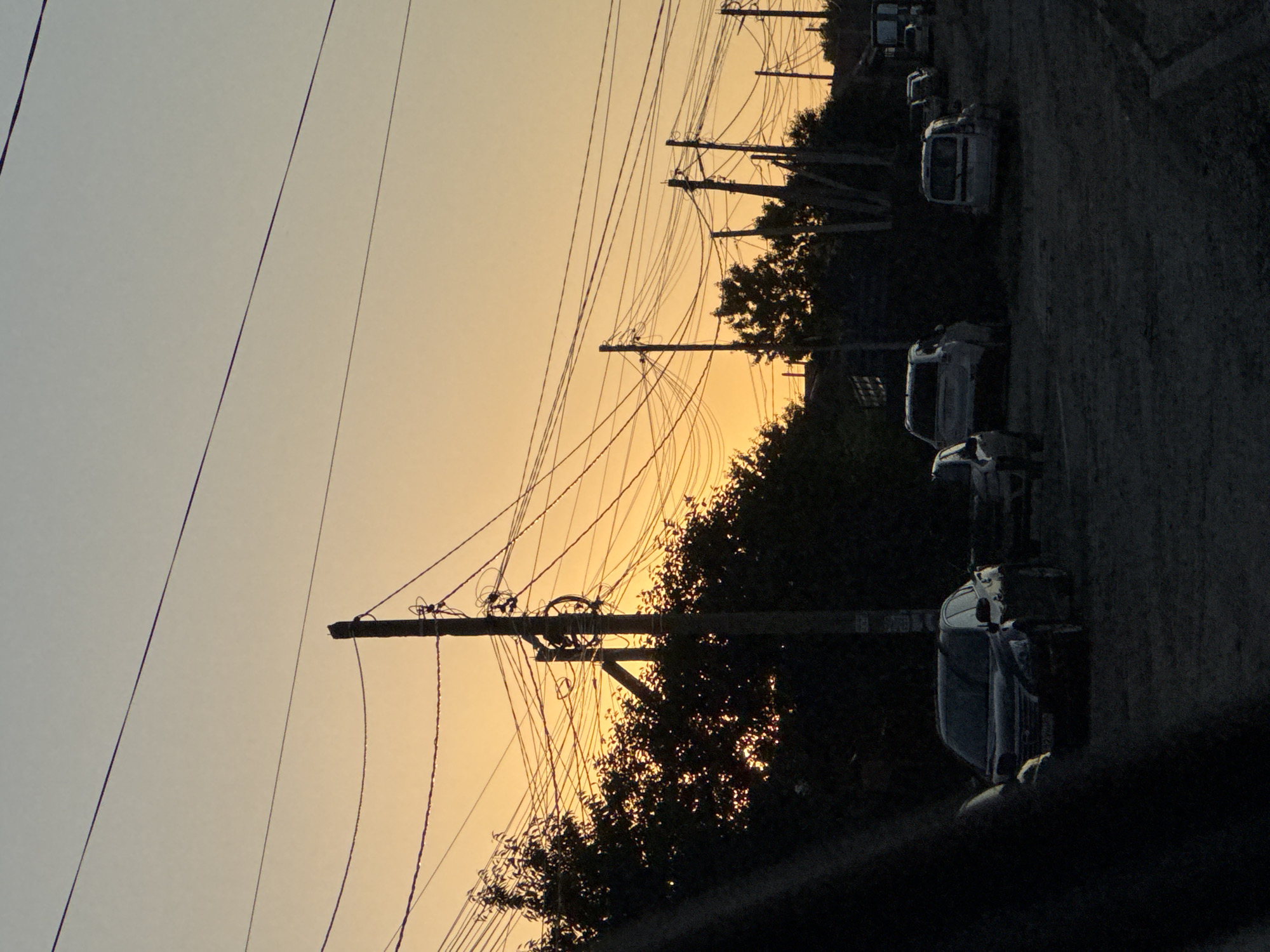 The image depicts a street scene in Mikhaylovsk, Russia, during what appears to be sunset or dawn due to the warm, orange hues in the sky. The orientation of the image is vertical. Overhead power lines crisscross the frame, supported by utility poles that are silhouetted against the sky. In the background, several vehicles, including what look like vans and possibly a small truck, are parked along the side of a dark, unpaved road. Dense trees with dark foliage line the edge of the road, creating a stark contrast with the bright sky. The overall mood is quiet and somewhat somber, possibly due to the low light conditions and the stillness of the scene. There is no visible text in the image.