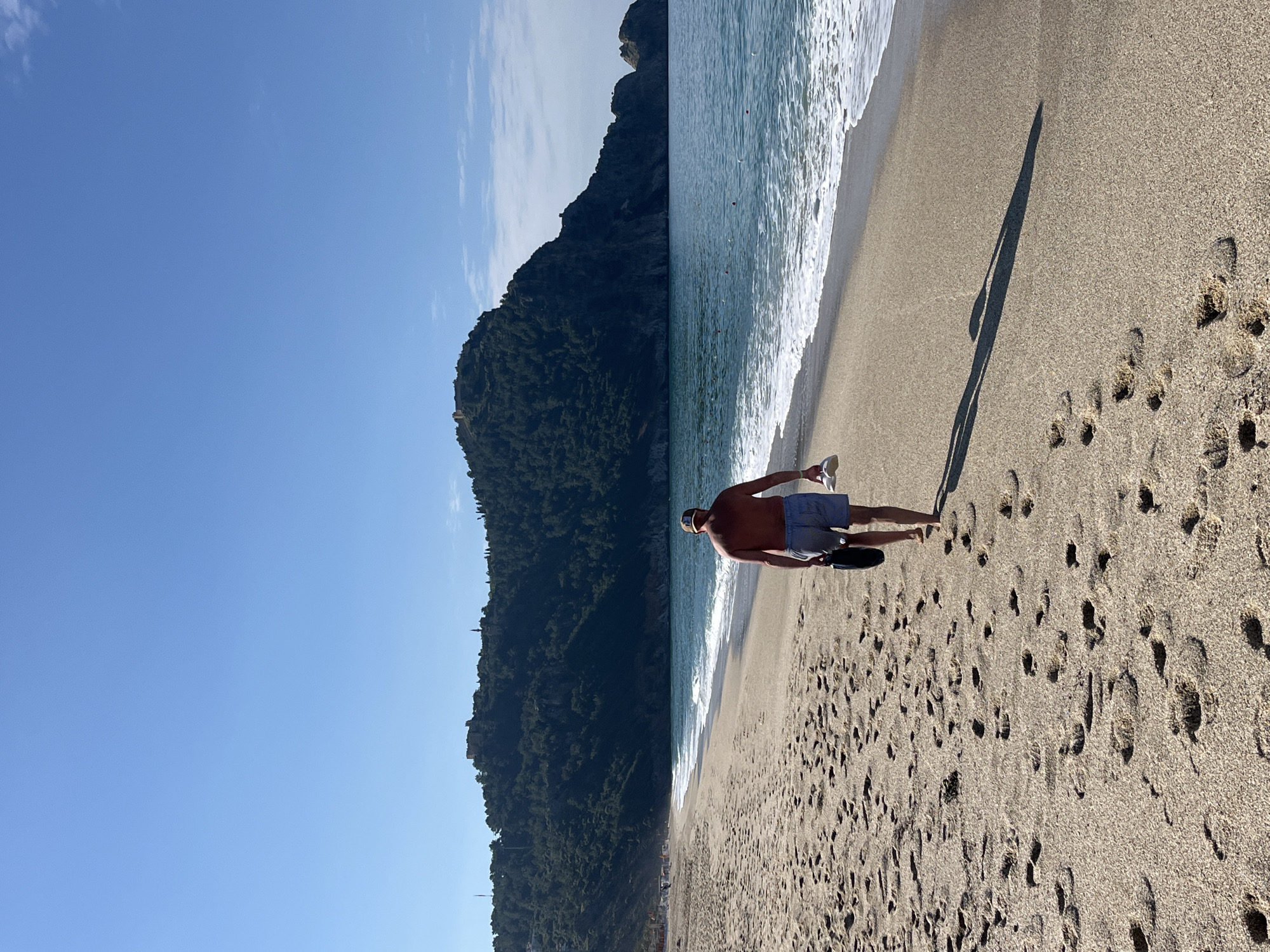 A medium shot captures a man walking along a beach. He is bare-chested, wearing shorts, a visor, and carrying footwear. The beach is sandy with numerous footprints. The edge of the ocean is marked by small waves. To the left, a verdant, hilly landscape meets the shoreline. A clear, blue sky is overhead. The scene is Alanya, Turkey.