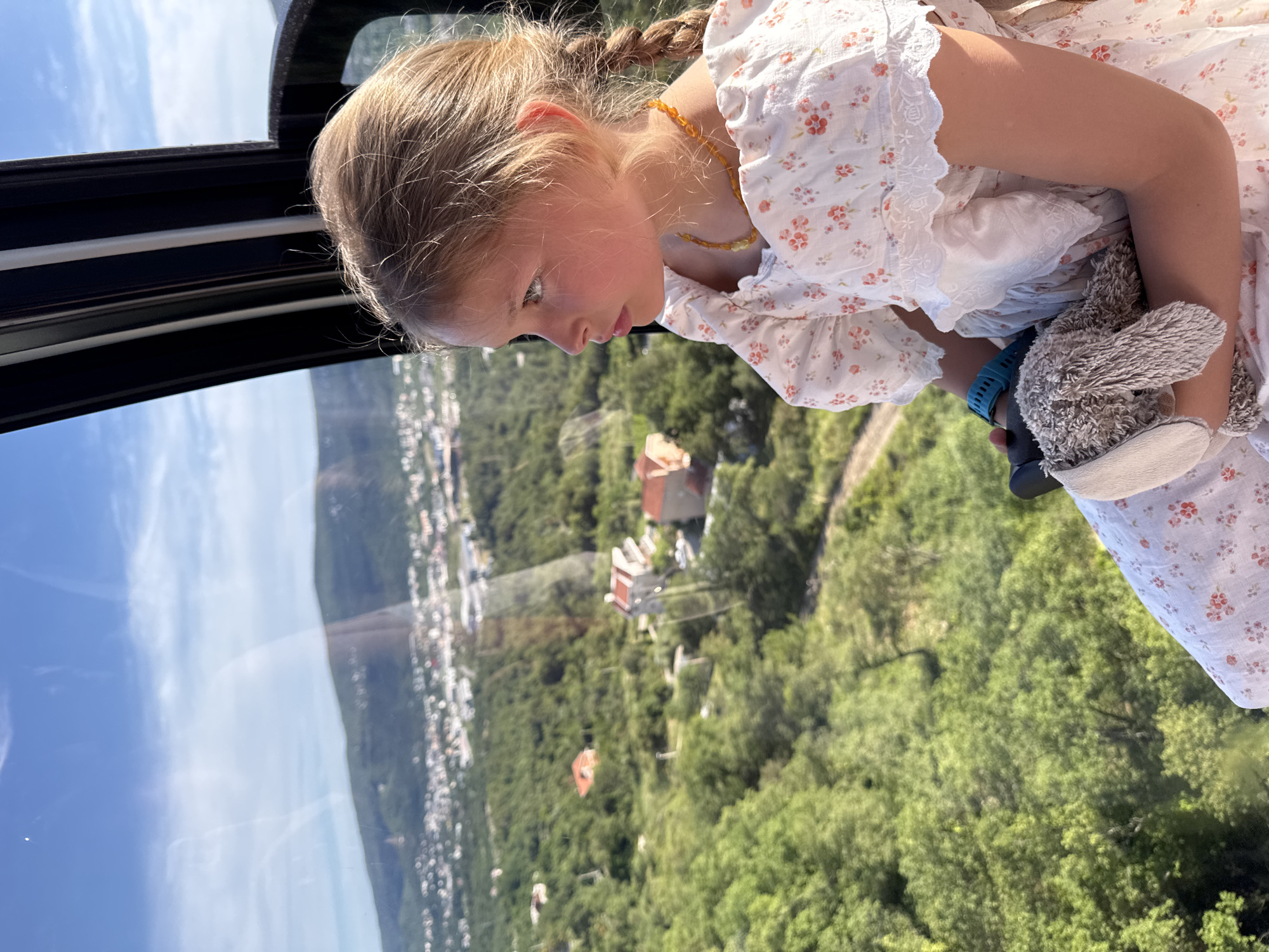 In the city of Dub, Montenegro, a young girl with blonde hair in braids looks out of the window of a cable car. She is wearing a white dress with a floral pattern and is holding a stuffed rabbit. The cable car is moving, offering a view of the city and the surrounding green hills. The sky is blue with scattered clouds, and the sun is shining, creating a bright, clear day. The scene appears to be a scenic tourist experience, with the cable car providing a unique perspective of the landscape.